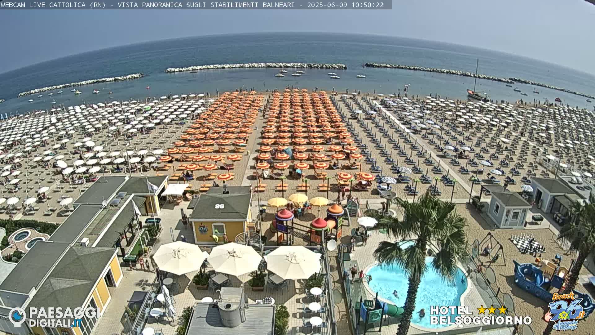 A sandy beach filled with numerous umbrellas and beachgoers is shown under a sunny sky, next to a calm sea with boats visible in the distance.