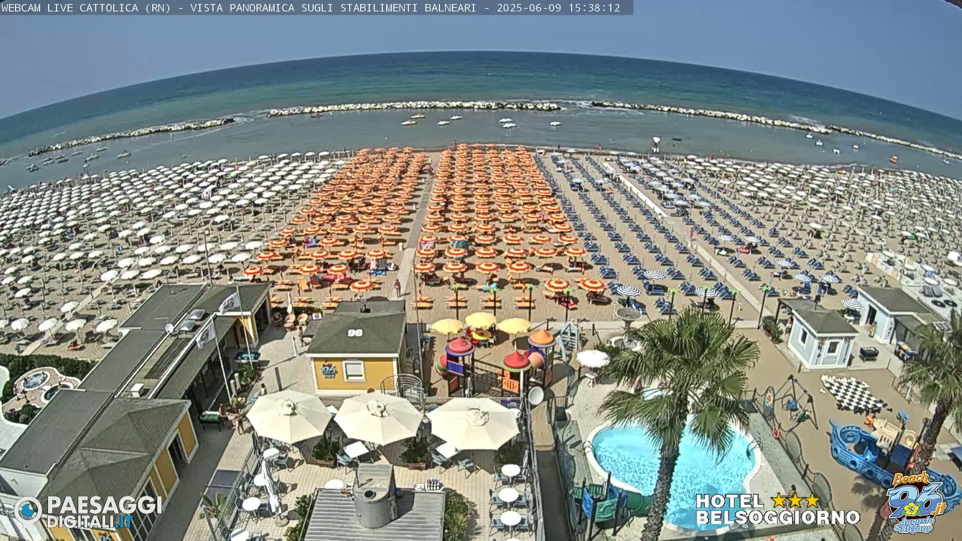 A crowded beach with numerous umbrellas and beach chairs under a sunny sky, adjacent to a hotel with a pool and playground.