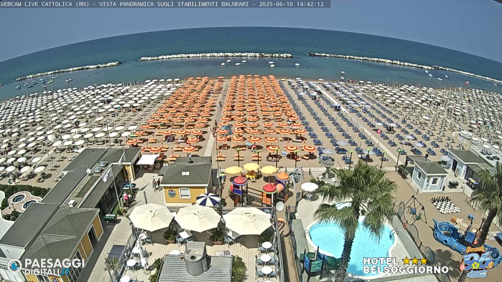 A sunny beach scene shows numerous rows of beach umbrellas and lounge chairs, a swimming pool, a playground, and a calm ocean.