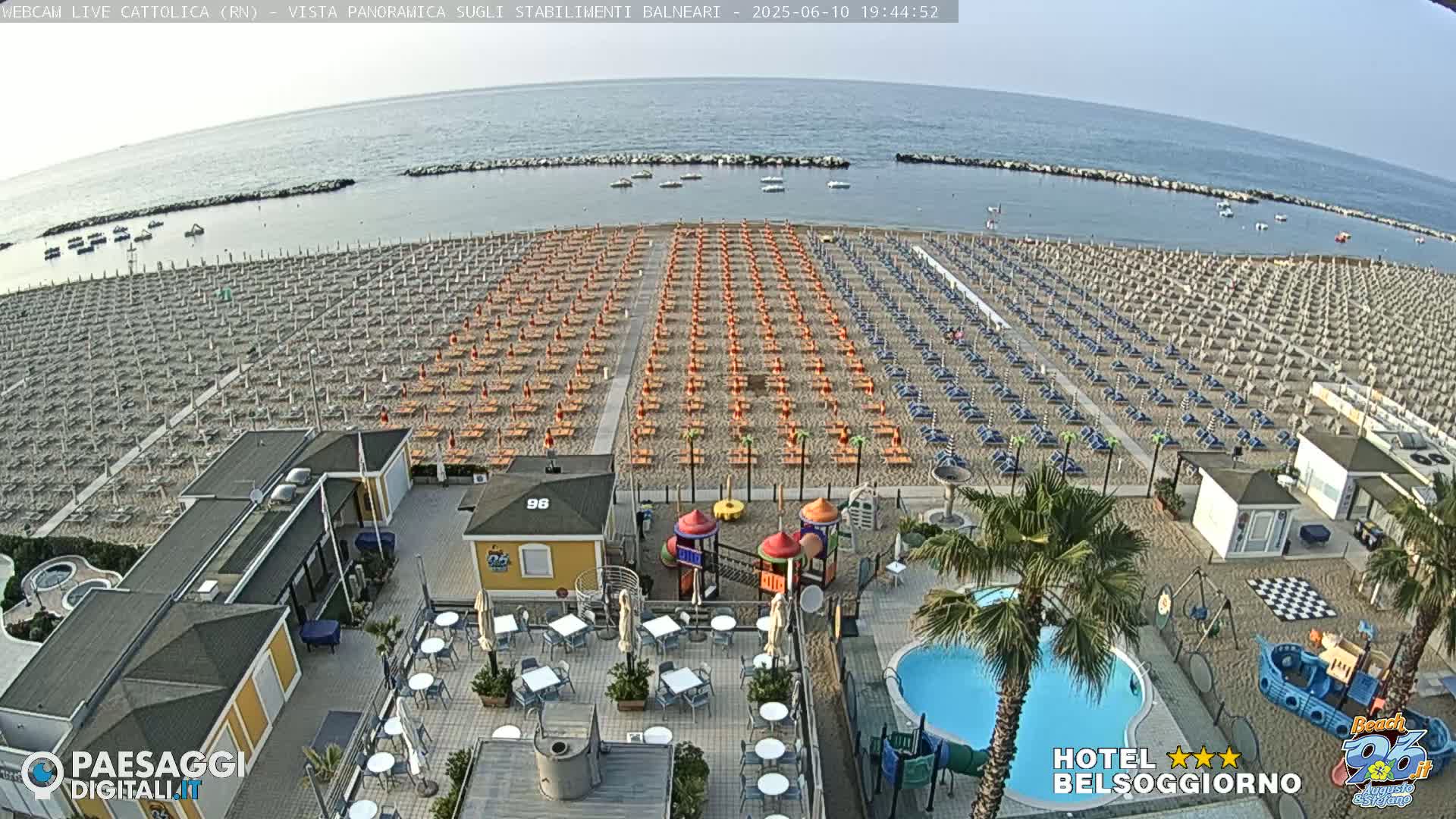 An aerial view of a beach with numerous umbrellas and lounge chairs, a small playground, a swimming pool, and a beachfront restaurant, on a clear day.