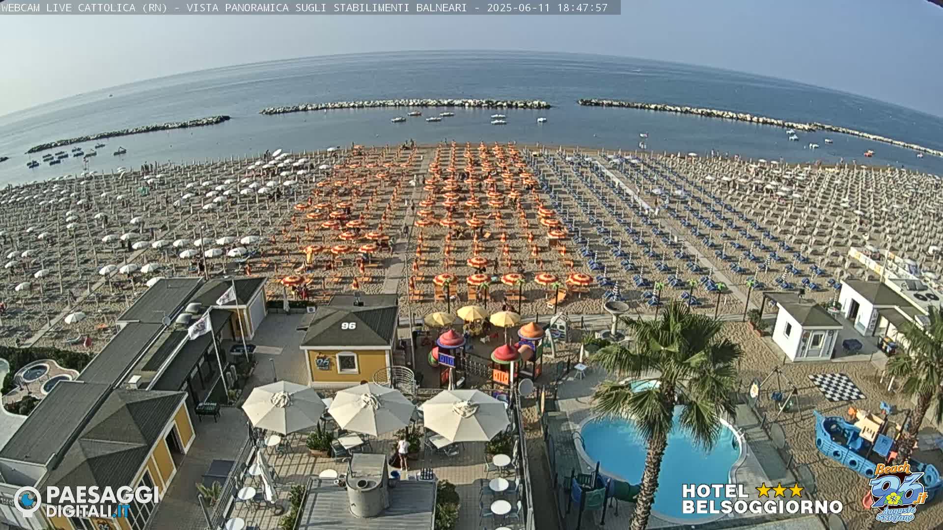 A crowded beach with rows of colorful umbrellas and beach chairs under a clear sky, overlooking a calm sea with a small playground and a swimming pool nearby.