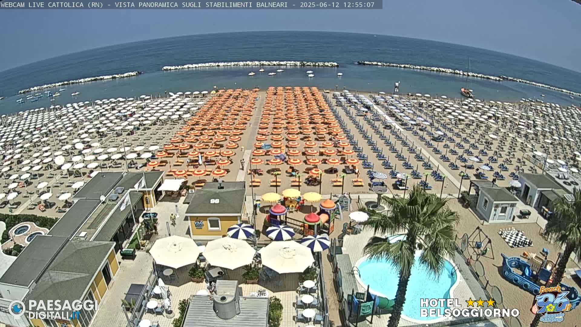 A sunny beach with numerous rows of umbrellas and lounge chairs, a swimming pool, and a playground is seen from an aerial view.