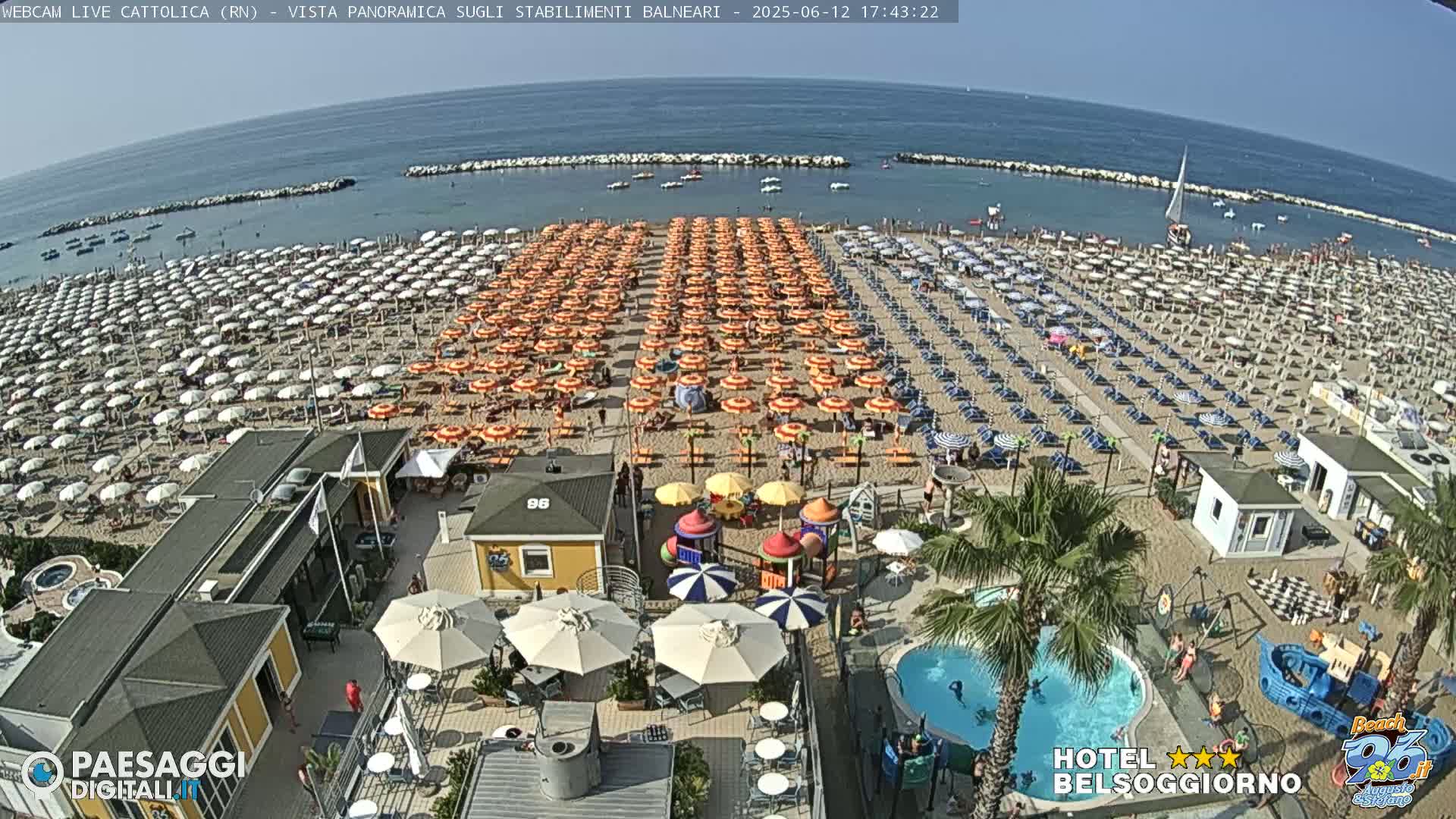 A crowded beach with numerous orange and white umbrellas, a children's play area, a small pool, and a calm ocean under a sunny sky.
