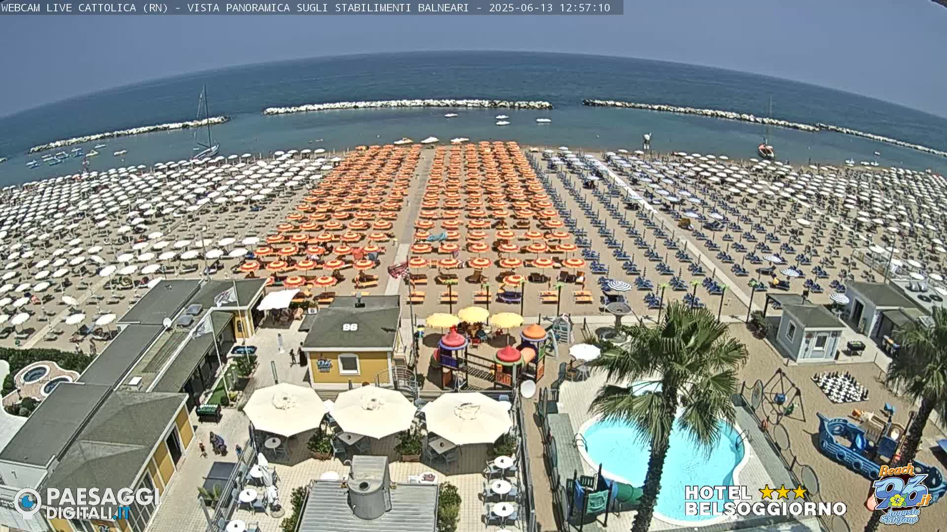A sunny beach scene shows rows of beach umbrellas and lounge chairs on a sandy beach next to a calm ocean, with a small pool and playground visible closer to the camera.