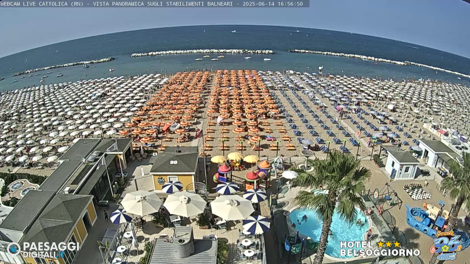 A sunny beach crowded with orange and white umbrellas, beachgoers, and beachside amenities including a small pool and playground.