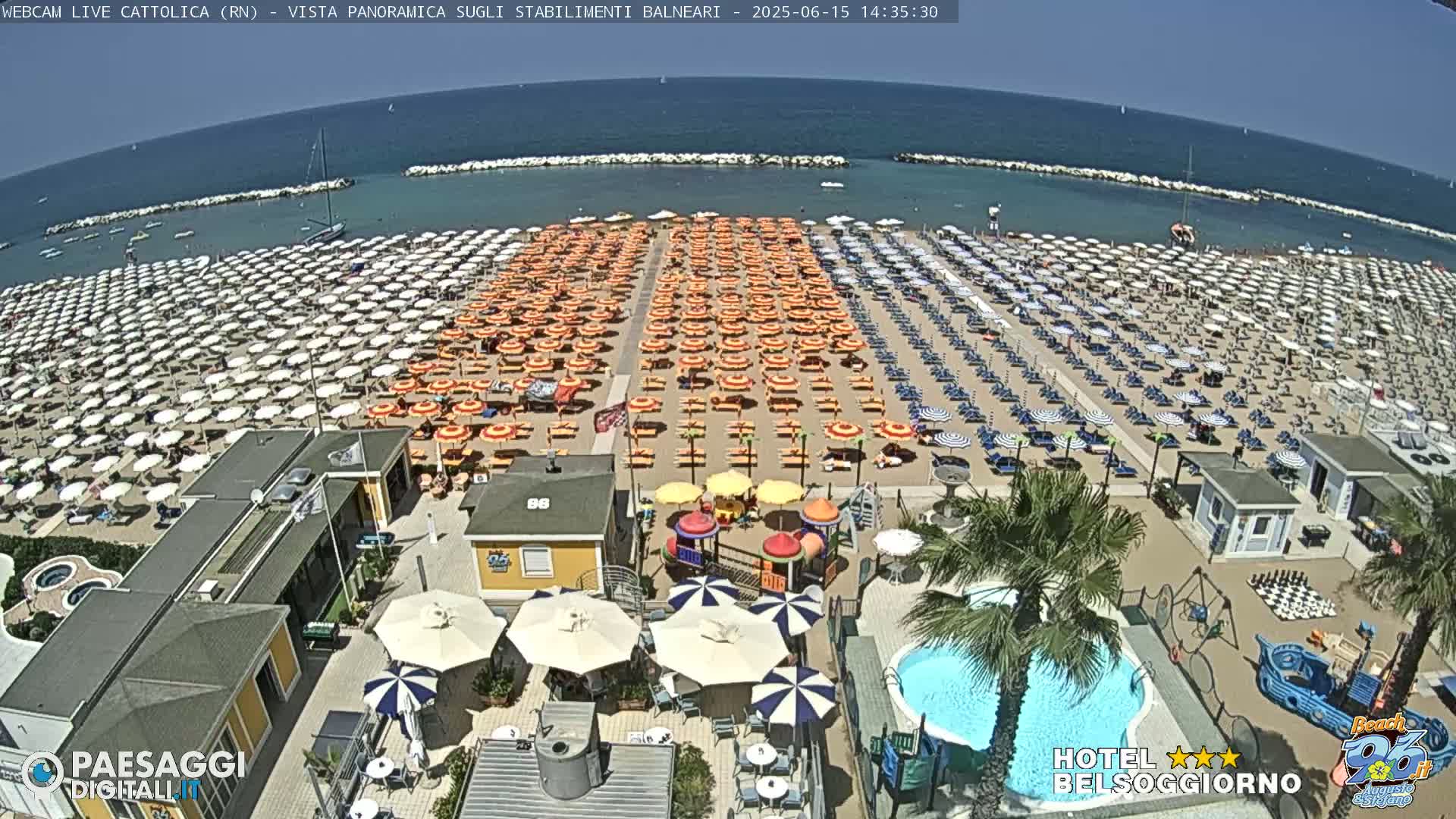 A sunny beach with numerous rows of beach umbrellas and lounge chairs, a small playground, and a swimming pool are visible from an elevated vantage point.