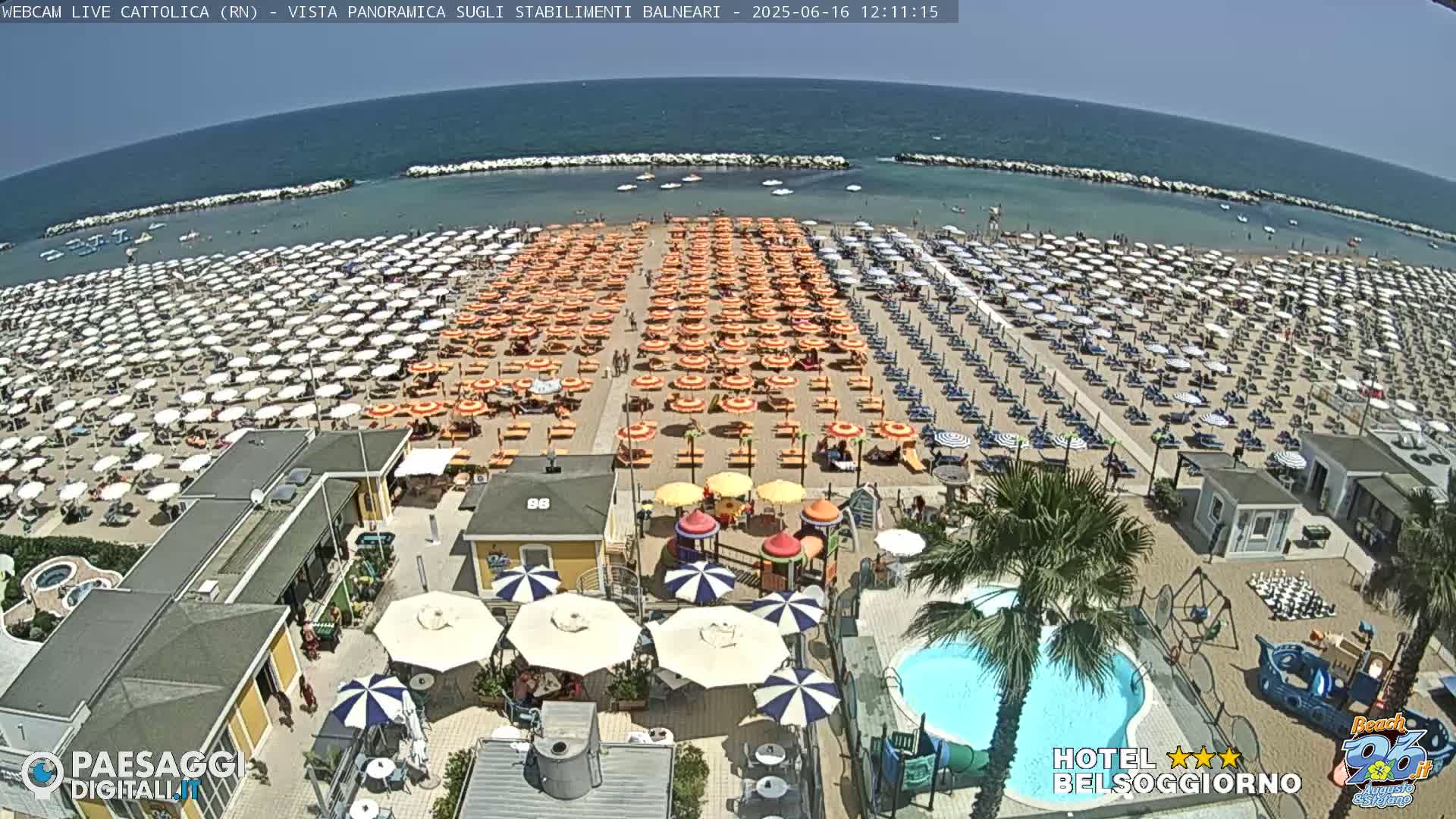 A crowded beach with numerous umbrellas and beach chairs under a sunny sky, adjacent to a swimming pool and playground.