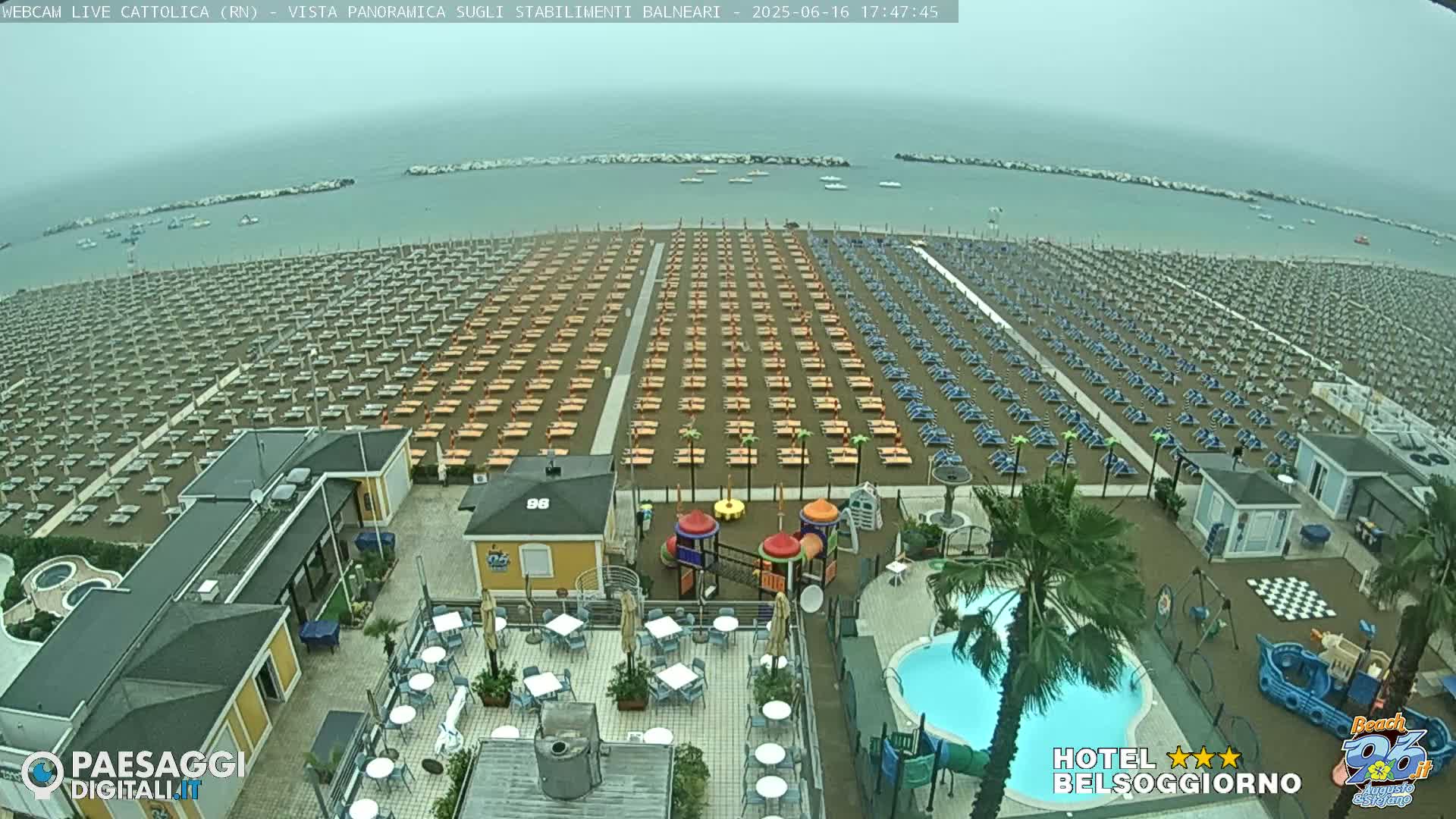 A beach with rows of empty beach umbrellas and lounge chairs is shown under an overcast sky, next to a swimming pool and playground, with a calm sea in the background.
