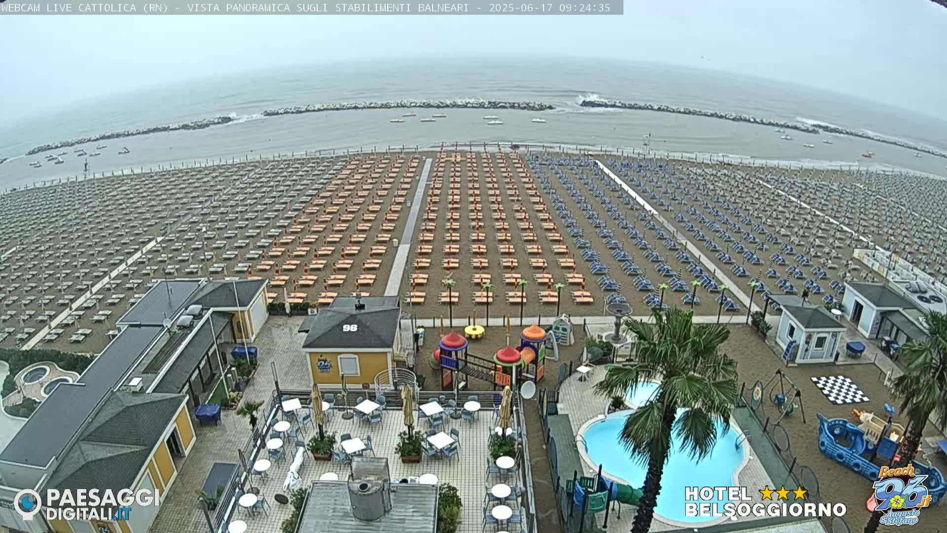 An overcast day reveals a beach lined with rows of unoccupied lounge chairs, adjacent to a resort with a pool and playground.