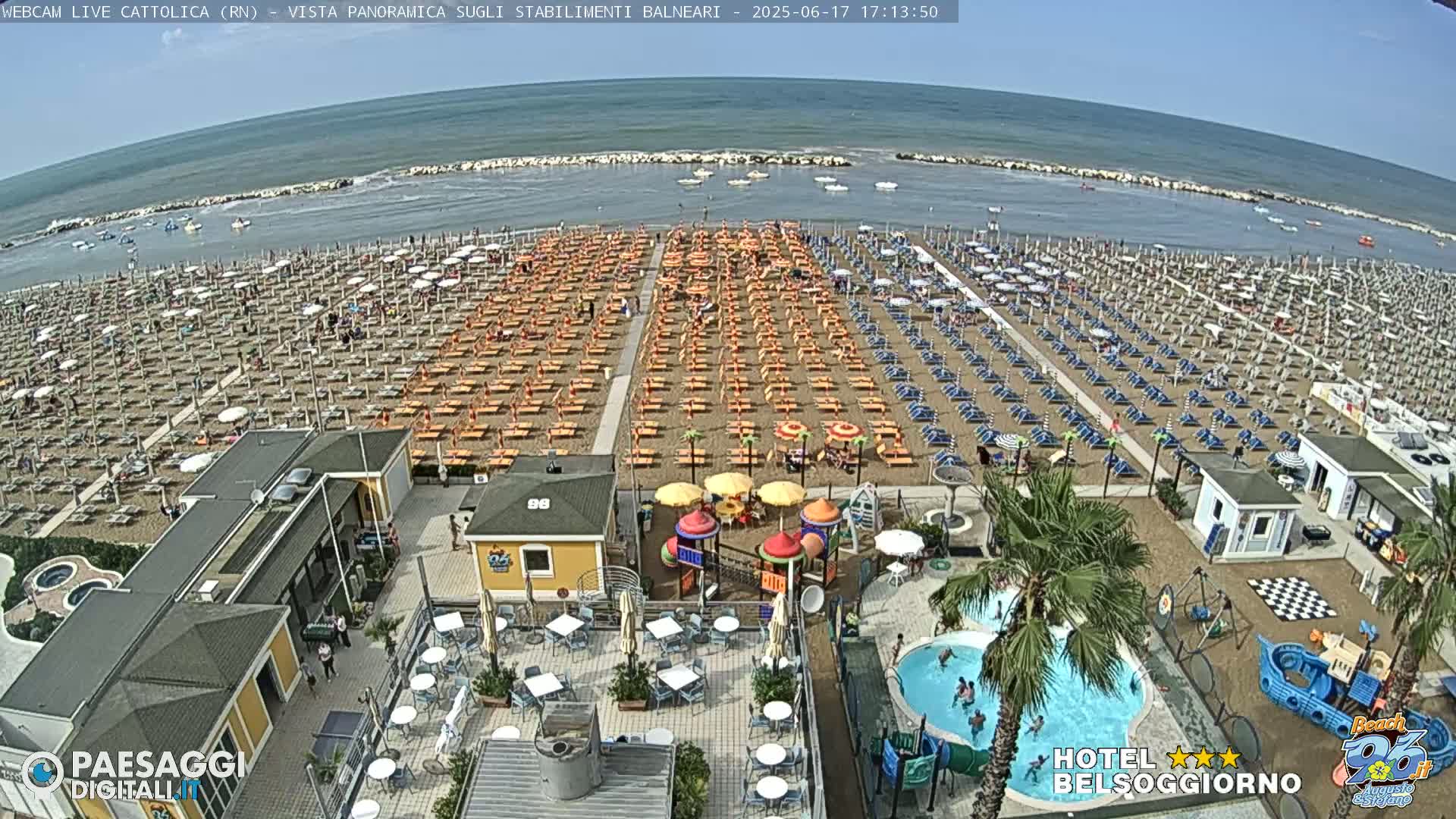 A crowded beach with rows of umbrellas and beach chairs, a playground, a pool, and several buildings under a sunny sky.