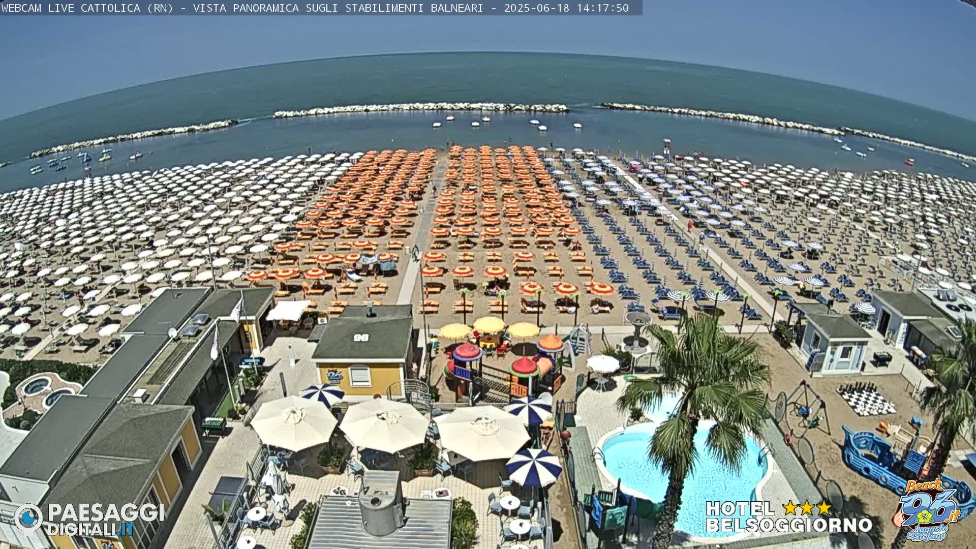 A beach with hundreds of umbrellas and lounge chairs under a bright sunny sky, adjacent to beachside buildings with a small pool and playground.