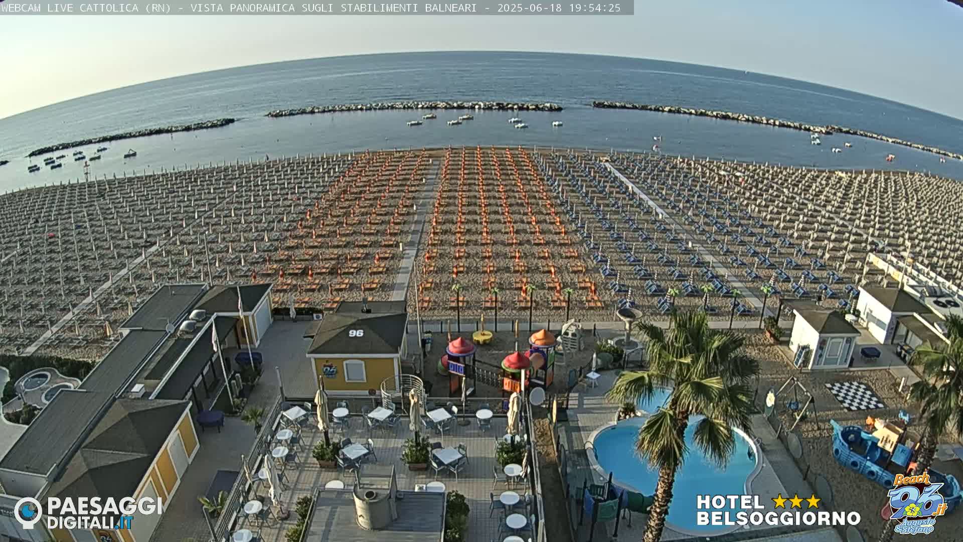 An aerial view of a beach shows numerous rows of beach umbrellas and lounge chairs, a small playground, a swimming pool, and calm ocean water under a clear sky.