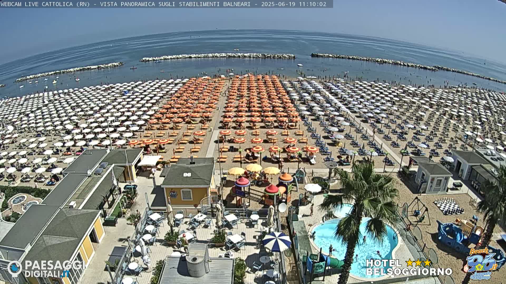 A beach with hundreds of umbrellas and beach chairs under a sunny sky, adjacent to a beachside establishment with a swimming pool and playground.