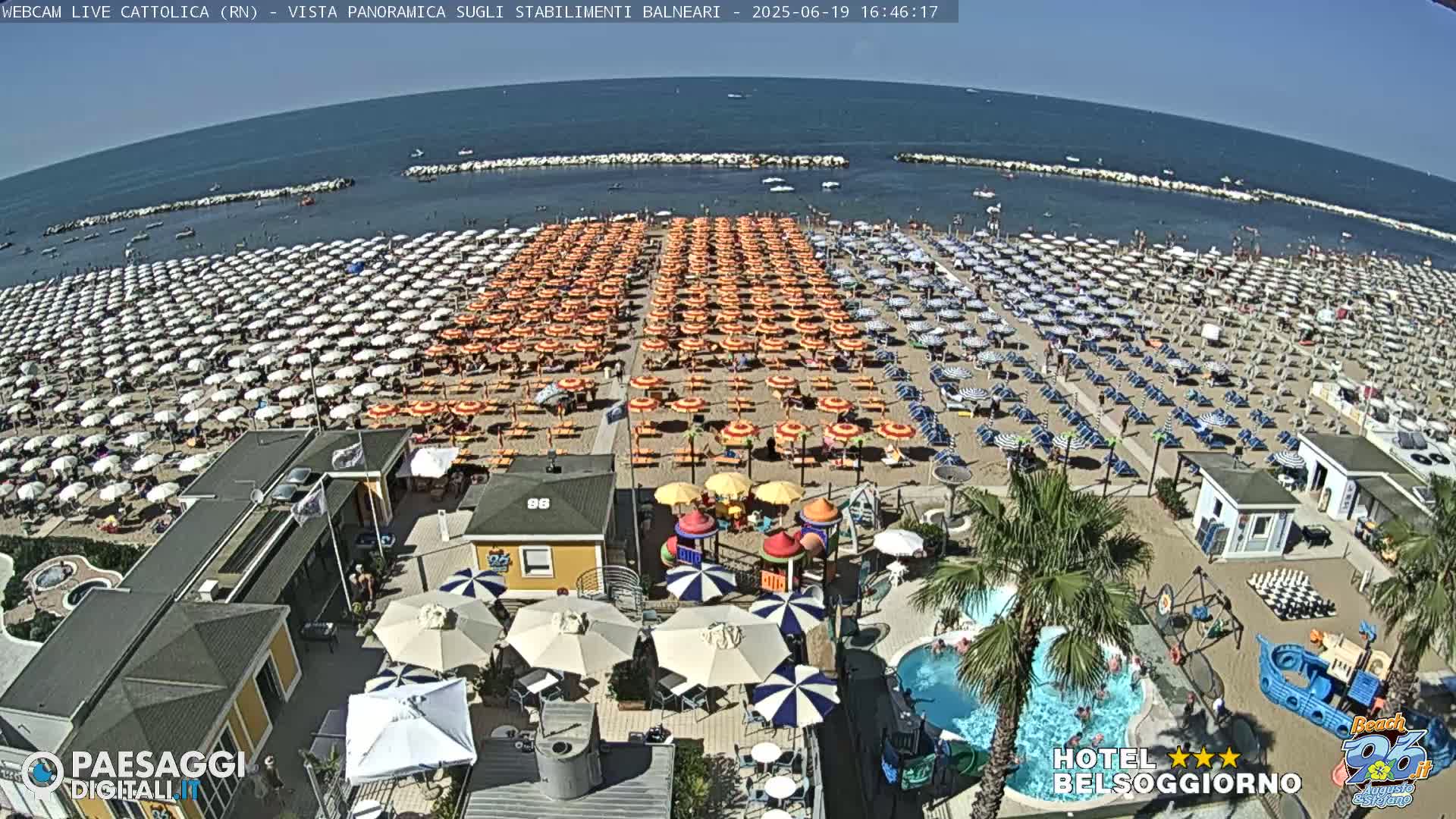 A crowded beach with numerous umbrellas and beachgoers under a sunny sky, adjacent to a beachfront establishment with a small pool and playground.