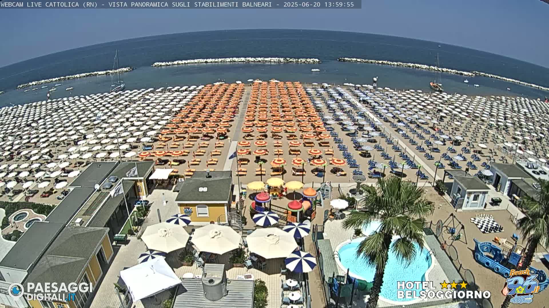 A beach with hundreds of umbrellas and lounge chairs under a clear, sunny sky, adjacent to a beachfront hotel with a pool and playground.