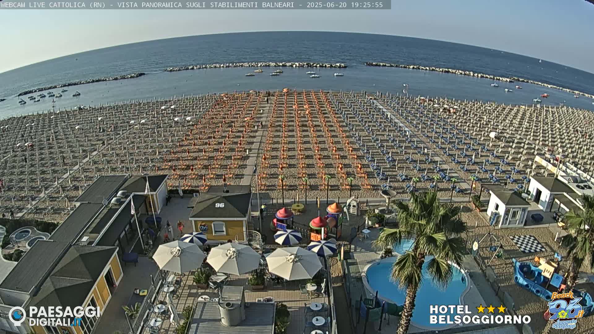 An aerial view shows a beach with numerous rows of umbrellas and lounge chairs, a small playground, a swimming pool, and several beachside buildings under a clear sky.