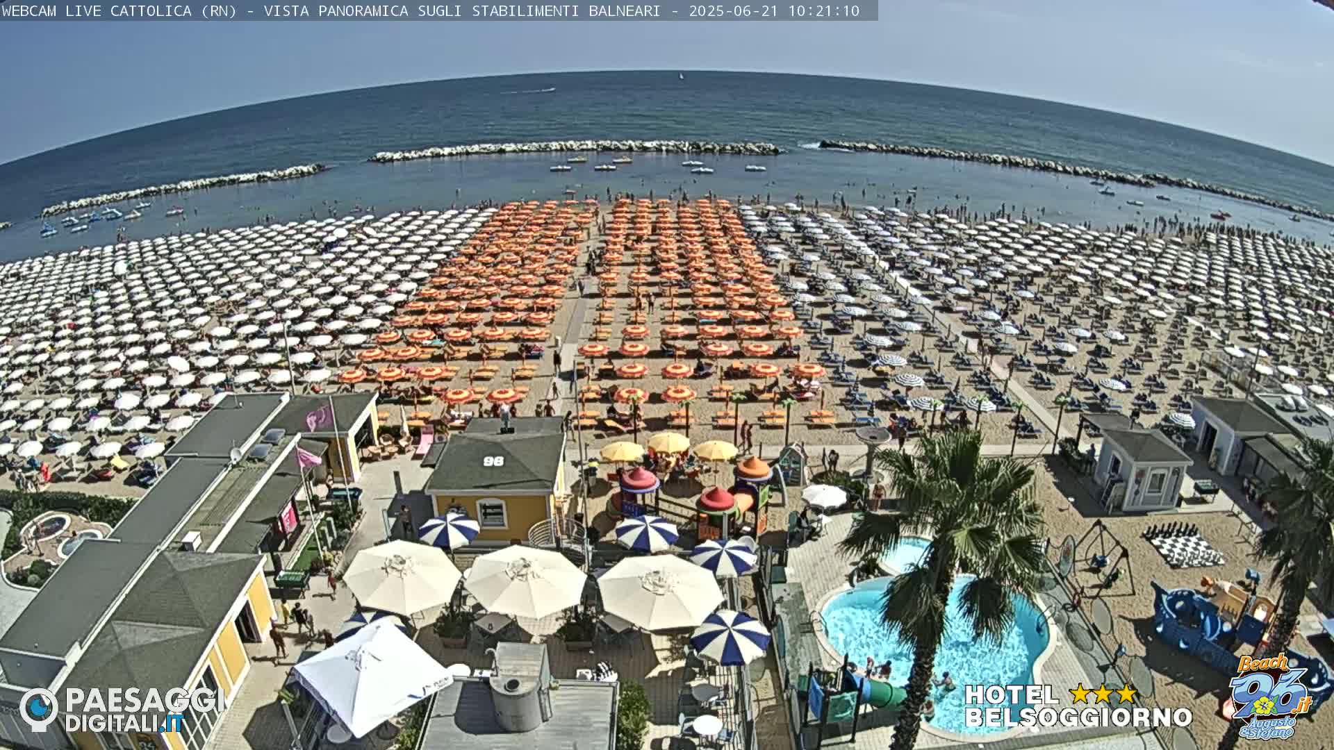 A crowded beach with numerous orange and white umbrellas under a clear blue sky, alongside a swimming pool and playground.