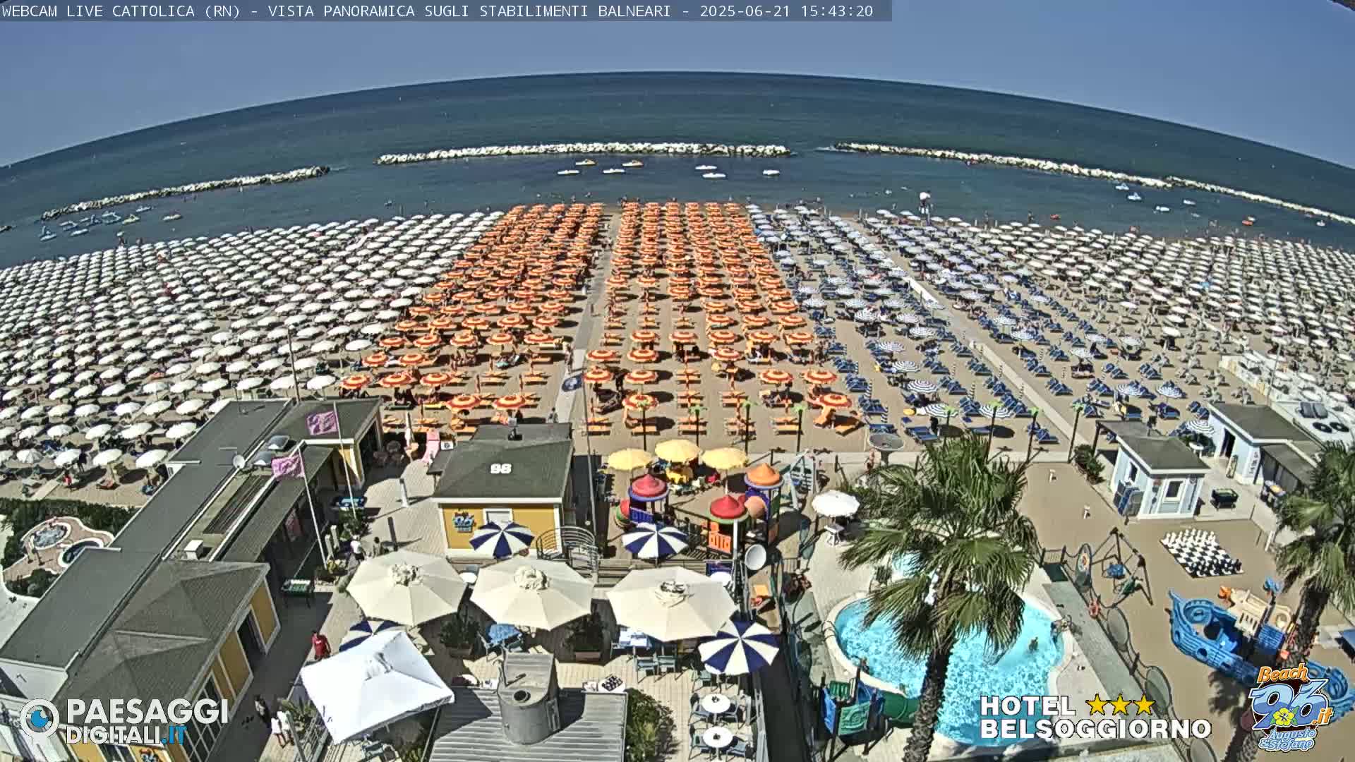 A sunny beach crowded with numerous orange and white umbrellas, adjacent to a beachside establishment with a small pool and playground.