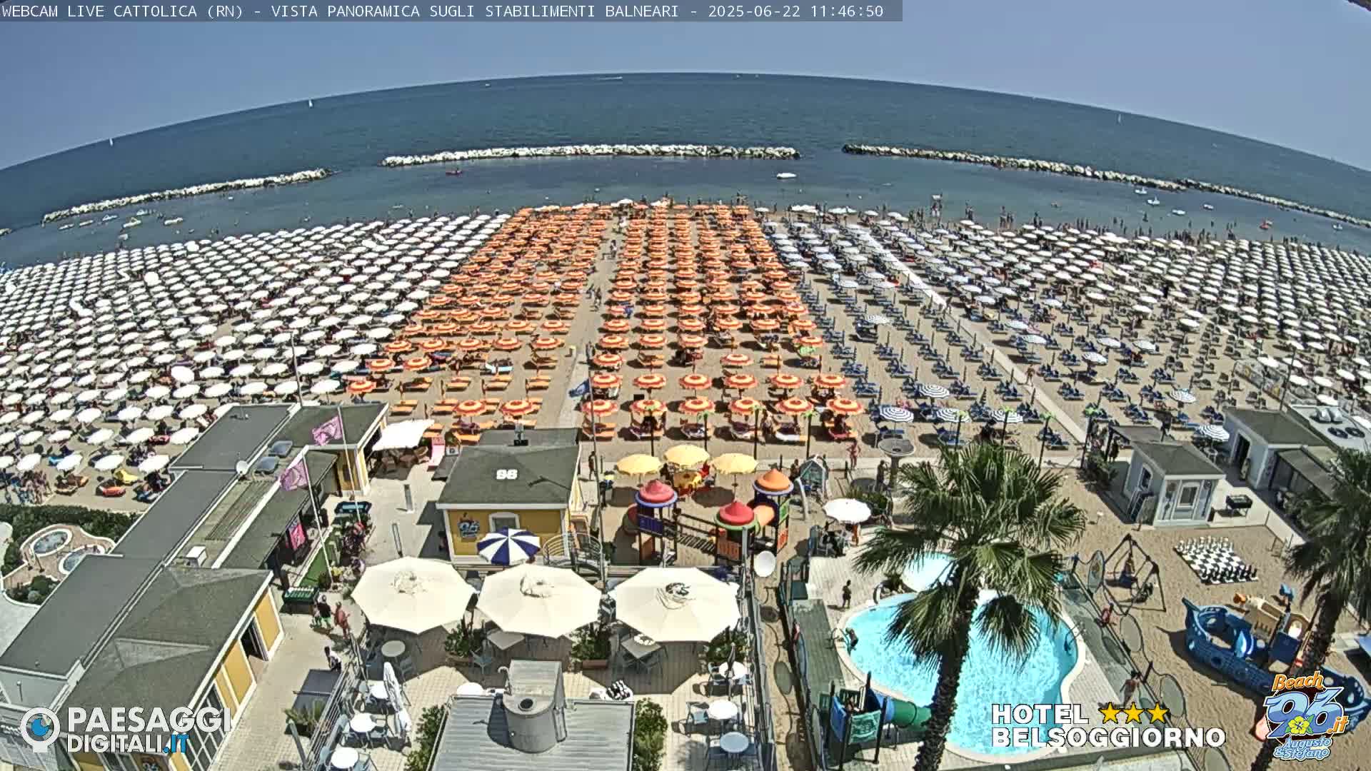 A crowded beach with numerous orange and white umbrellas under a sunny sky, adjacent to a swimming pool and playground.