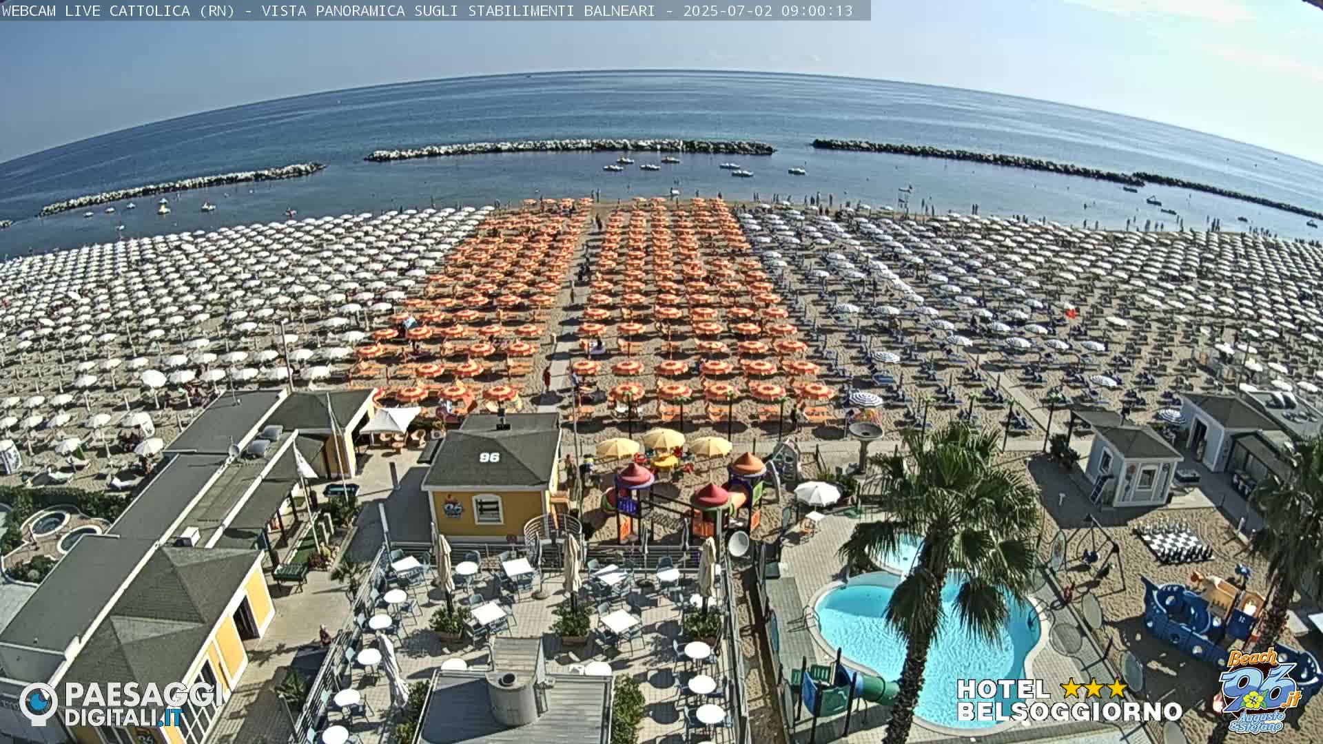 A beach with numerous rows of umbrellas and beach chairs, adjacent to a beachfront establishment with a pool and playground, on a sunny day.