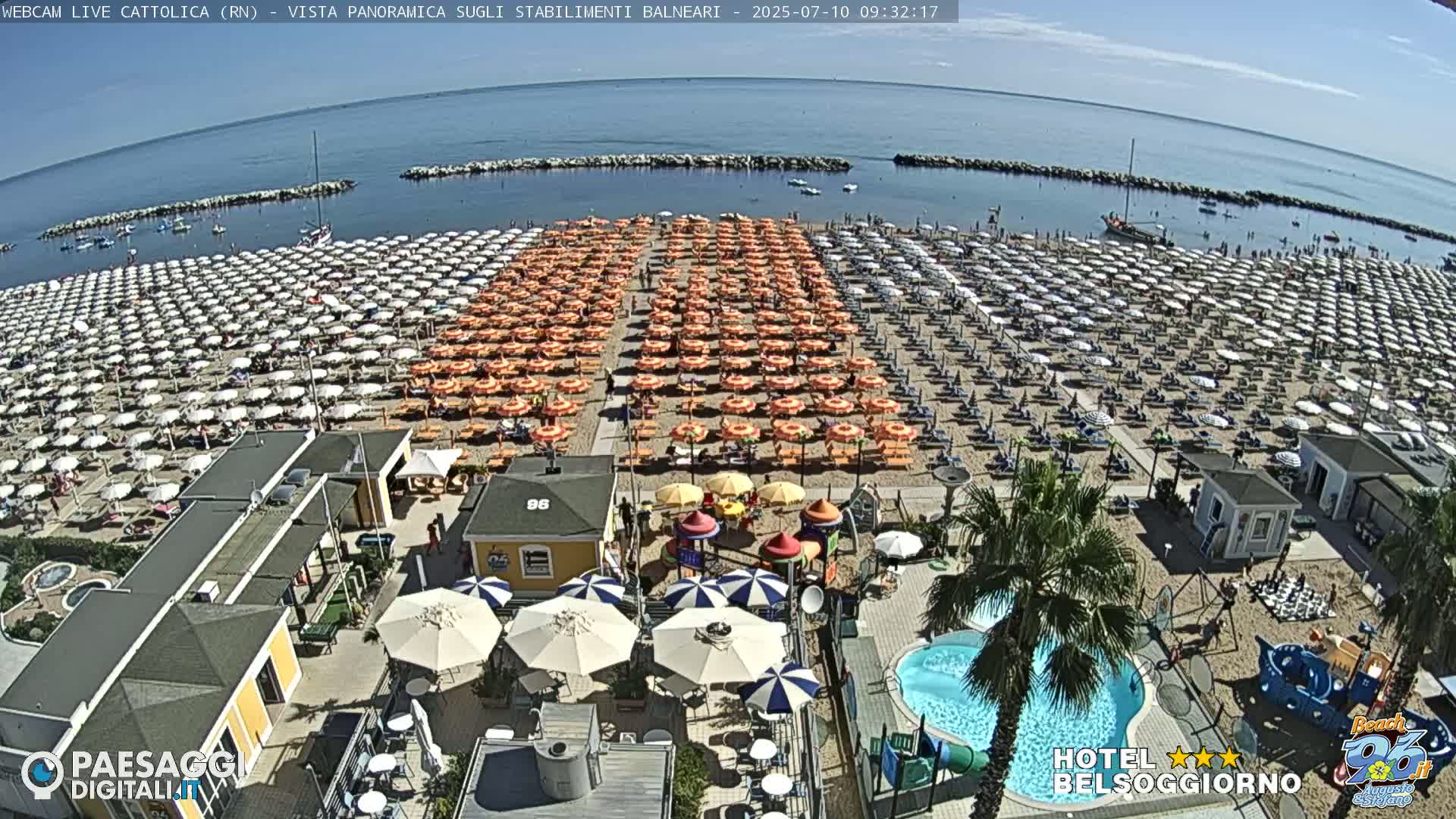 A beach crowded with numerous orange and white umbrellas, a small swimming pool, and a playground is seen on a sunny day, with calm blue water visible in the background.