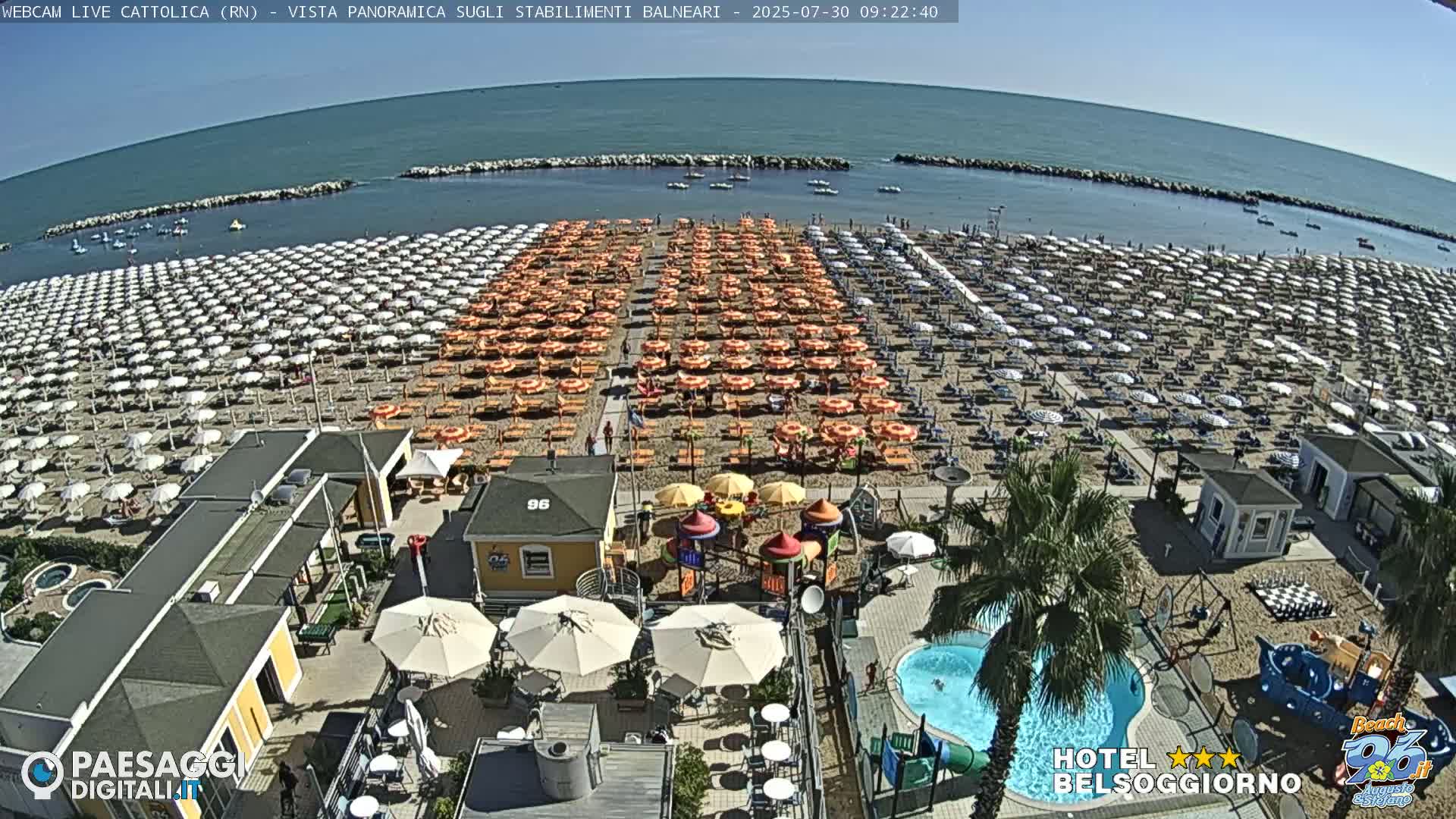 A sunny beach with numerous rows of umbrellas and beach chairs, a small playground, a swimming pool, and beachside buildings.
