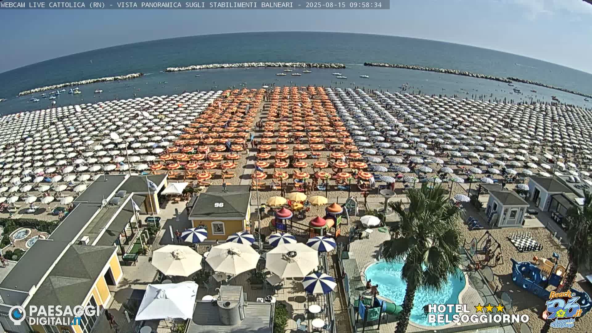 A sunny beach scene shows hundreds of beach umbrellas arranged in rows on the sand, with a small pool and playground visible closer to the camera.