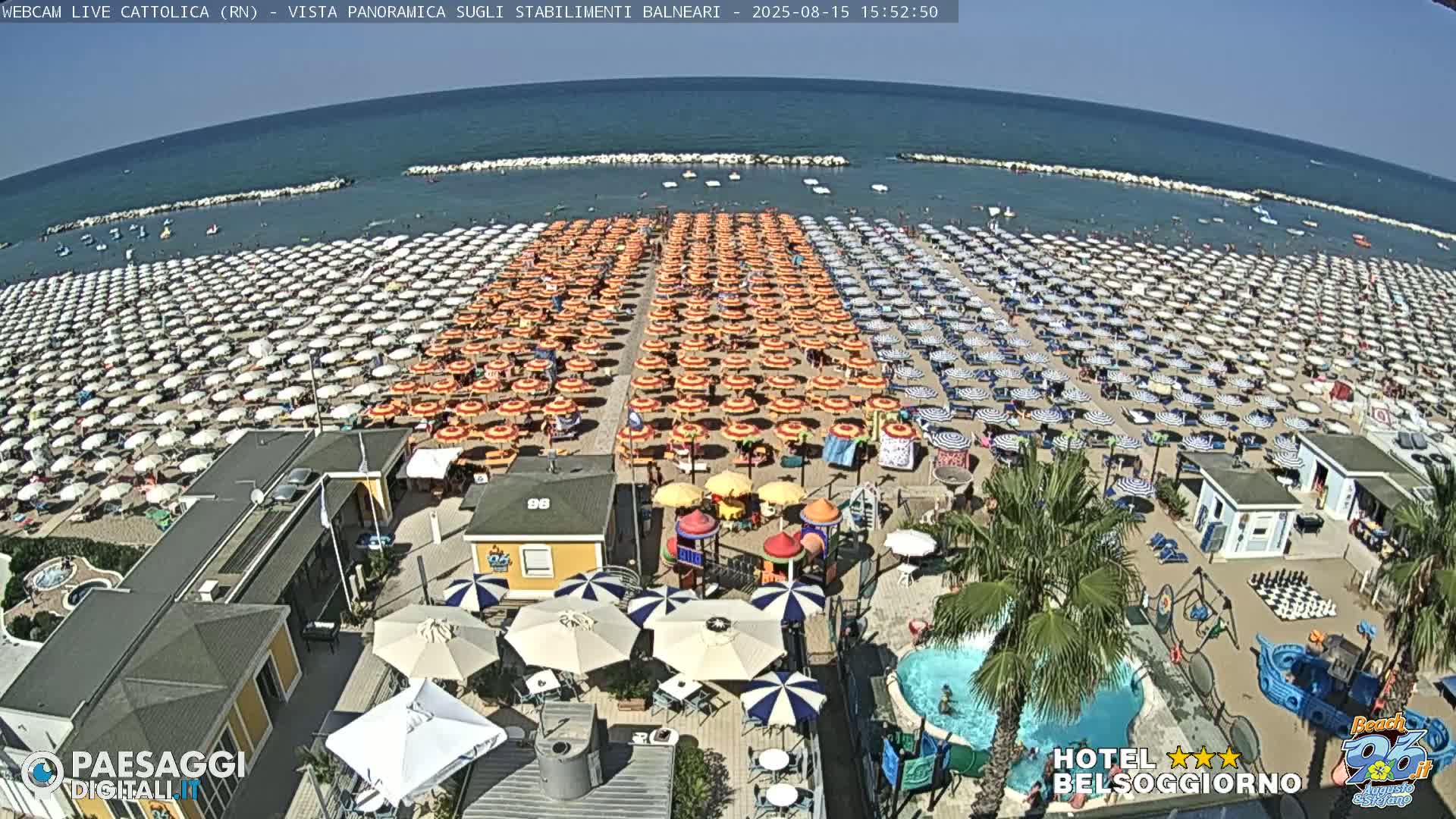 A sunny beach scene shows hundreds of beach umbrellas arranged in rows on the sand, with a small pool and playground visible closer to the camera.