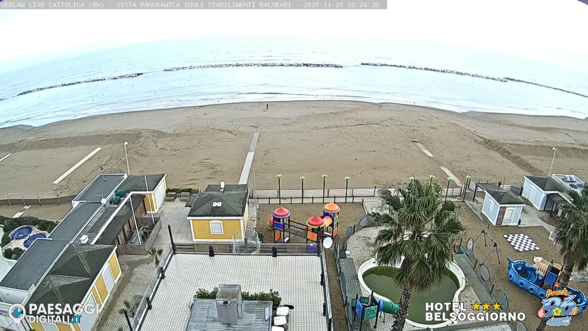 Under an overcast sky, a panoramic view showcases a wide sandy beach stretching towards an ocean with breakwaters, overlooked by a resort area featuring various buildings, colorful playgrounds, numerous palm trees, and a green-tinged pool.