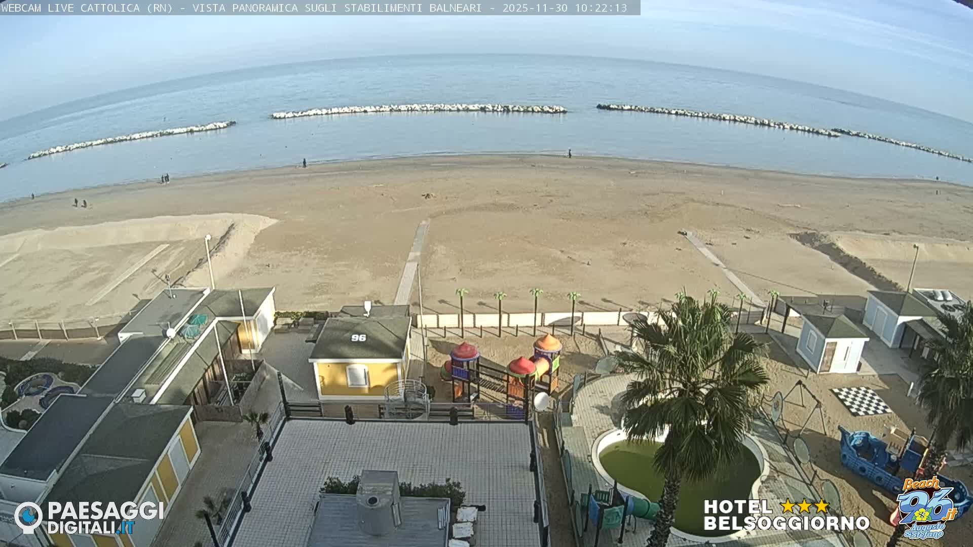 On a clear, sunny day, a wide sandy beach with a calm blue sea and rock breakwaters is overlooked by various beach buildings, palm trees, and children's play areas, including an empty pool.