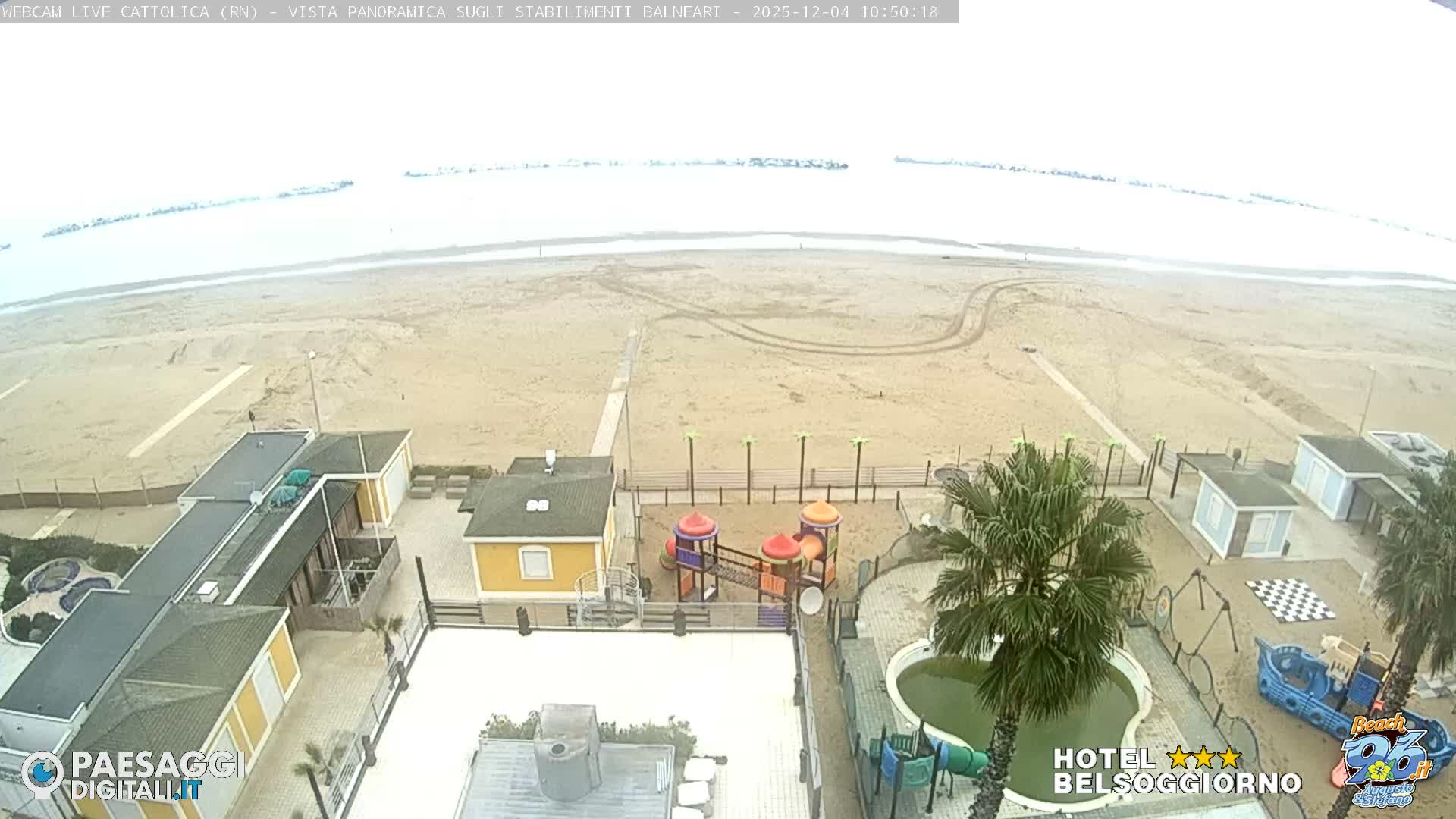 An aerial view captures a wide, sandy beach and a calm, light-colored sea under an overcast sky, with numerous beachside facilities including buildings, colorful playgrounds, a swimming pool, and palm trees in the foreground.