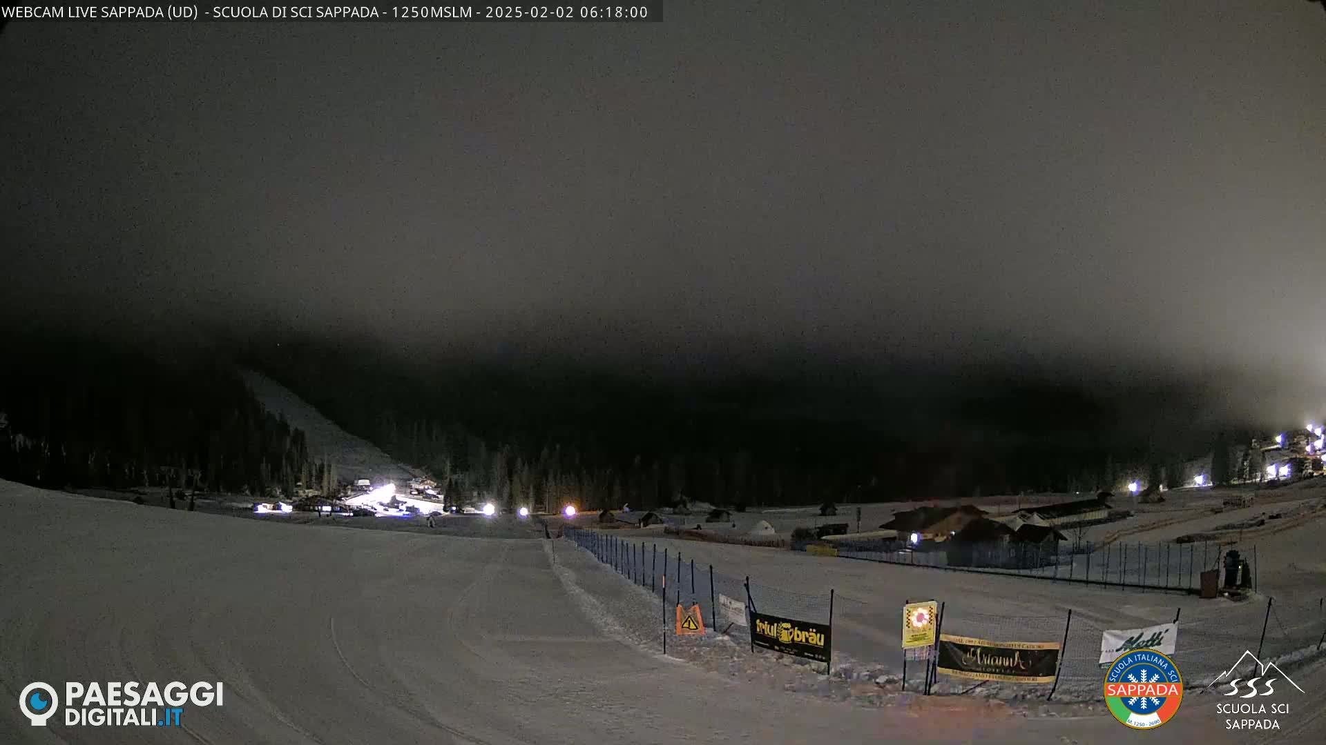 A nighttime view of a snow-covered ski slope with low fog, illuminated by lights from buildings and vehicles below.