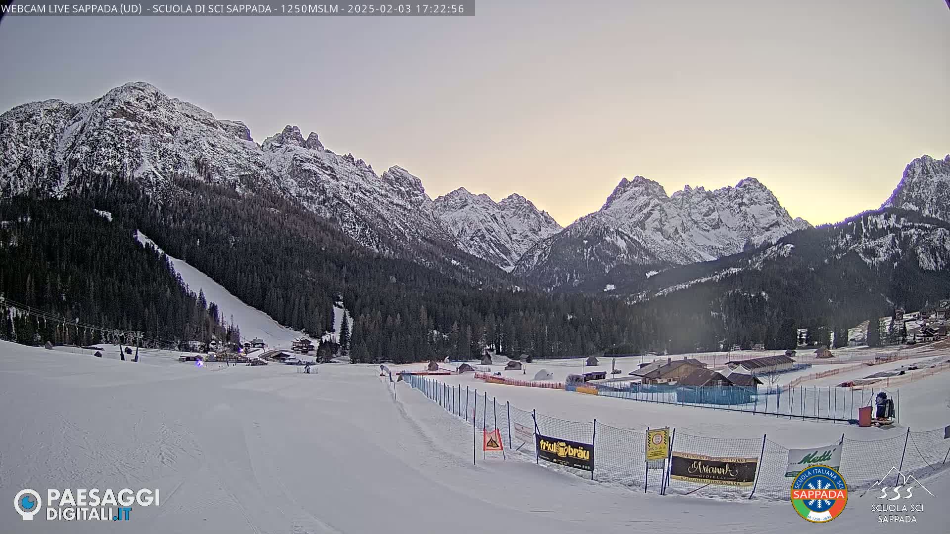 A snow-covered ski slope and resort are nestled in a valley between snow-capped mountains under a twilight sky.