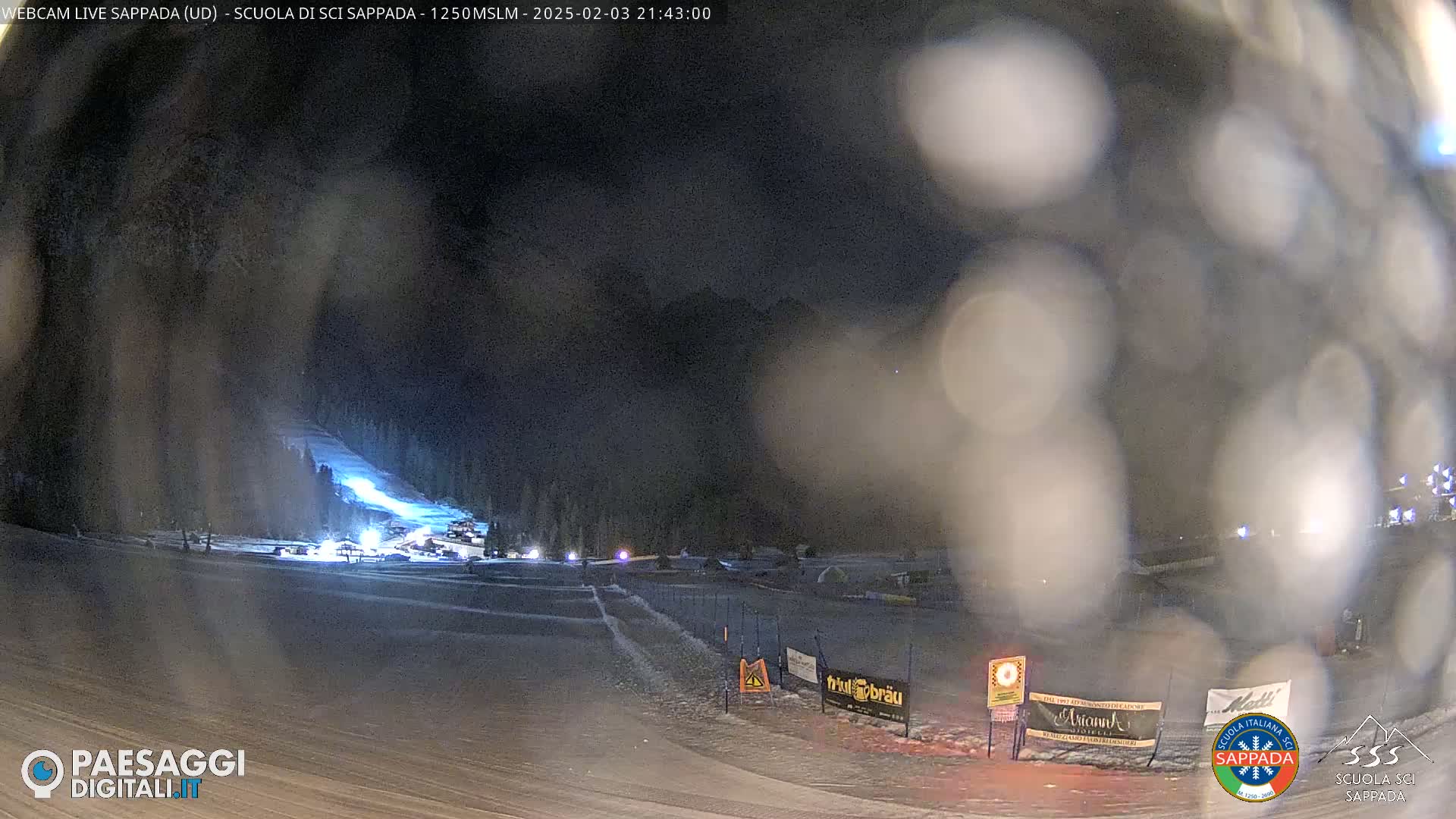 A nighttime view of a snow-covered ski slope with lights illuminating a section of the run, under a dark, possibly snowy sky.