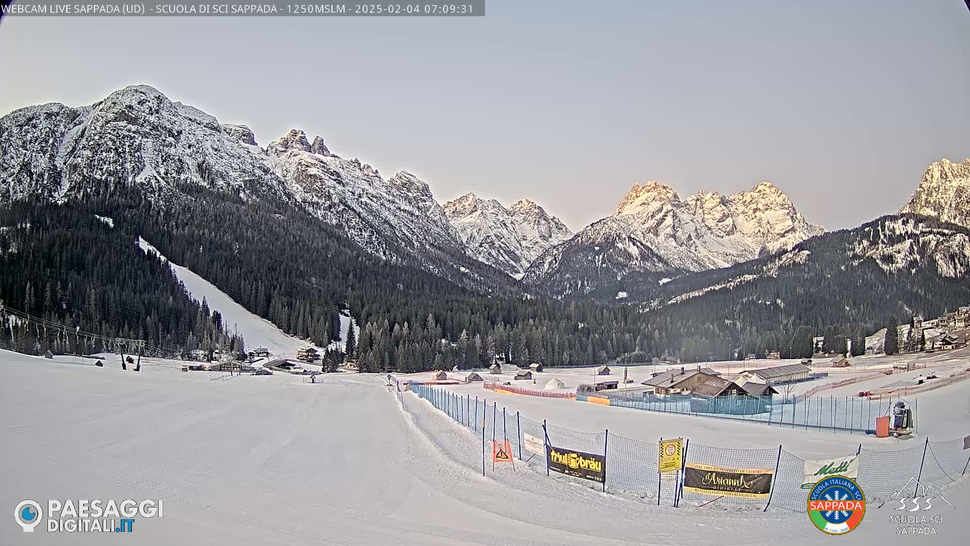 A snow-covered ski slope and surrounding forest are nestled beneath a range of snow-capped mountains under a clear, pale sky at dawn.