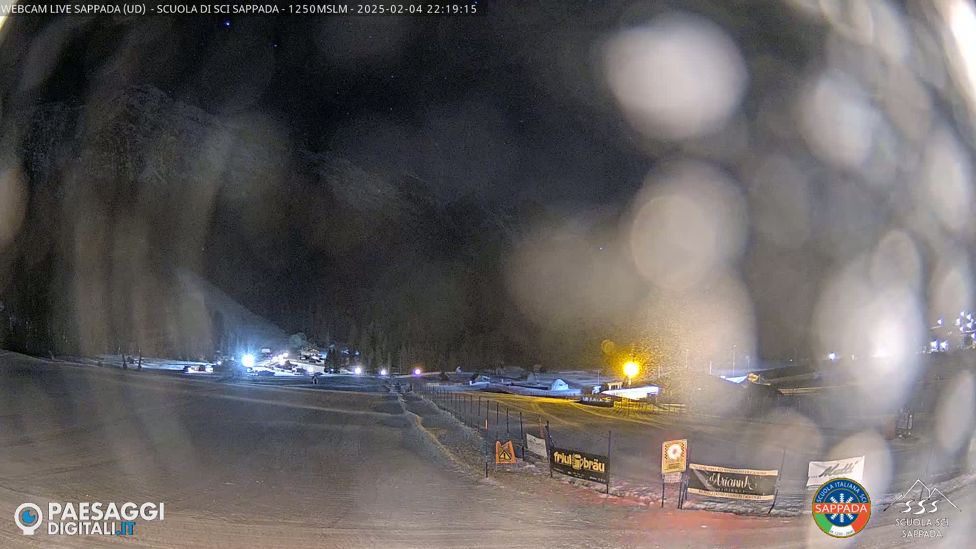 A nighttime snow scene shows a snow-covered slope with lights illuminating a ski area under falling snow.