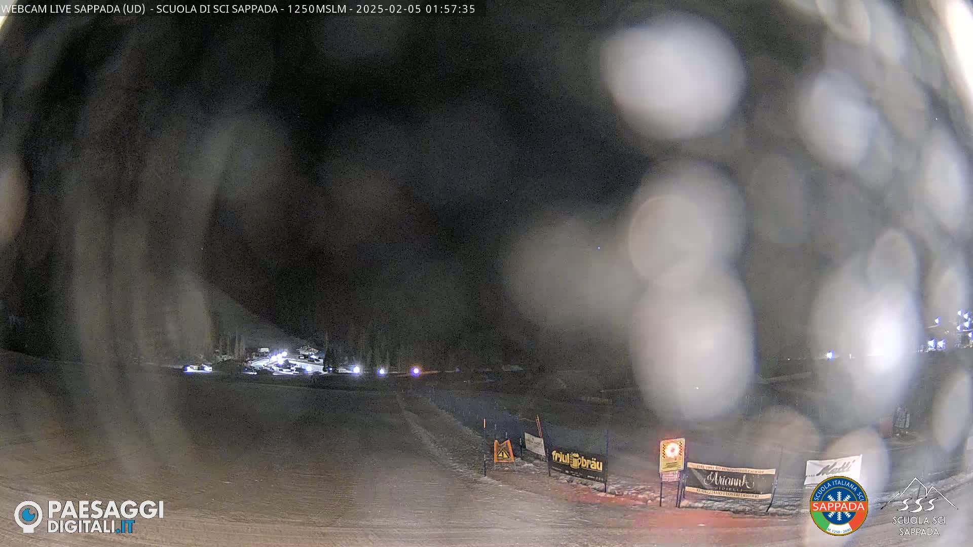 A nighttime, snowy scene shows a road with several cars and a few signs, viewed through a somewhat blurry lens.