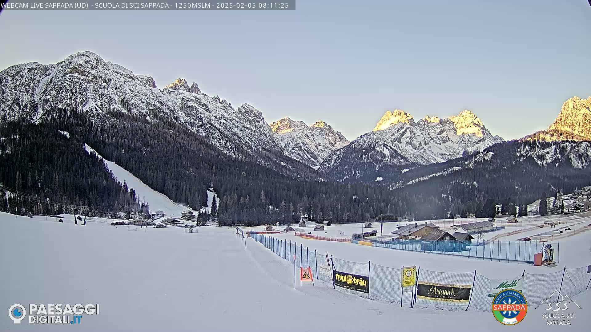 A snow-covered ski slope is situated in a valley below snow-capped mountains under a clear, bright sky.