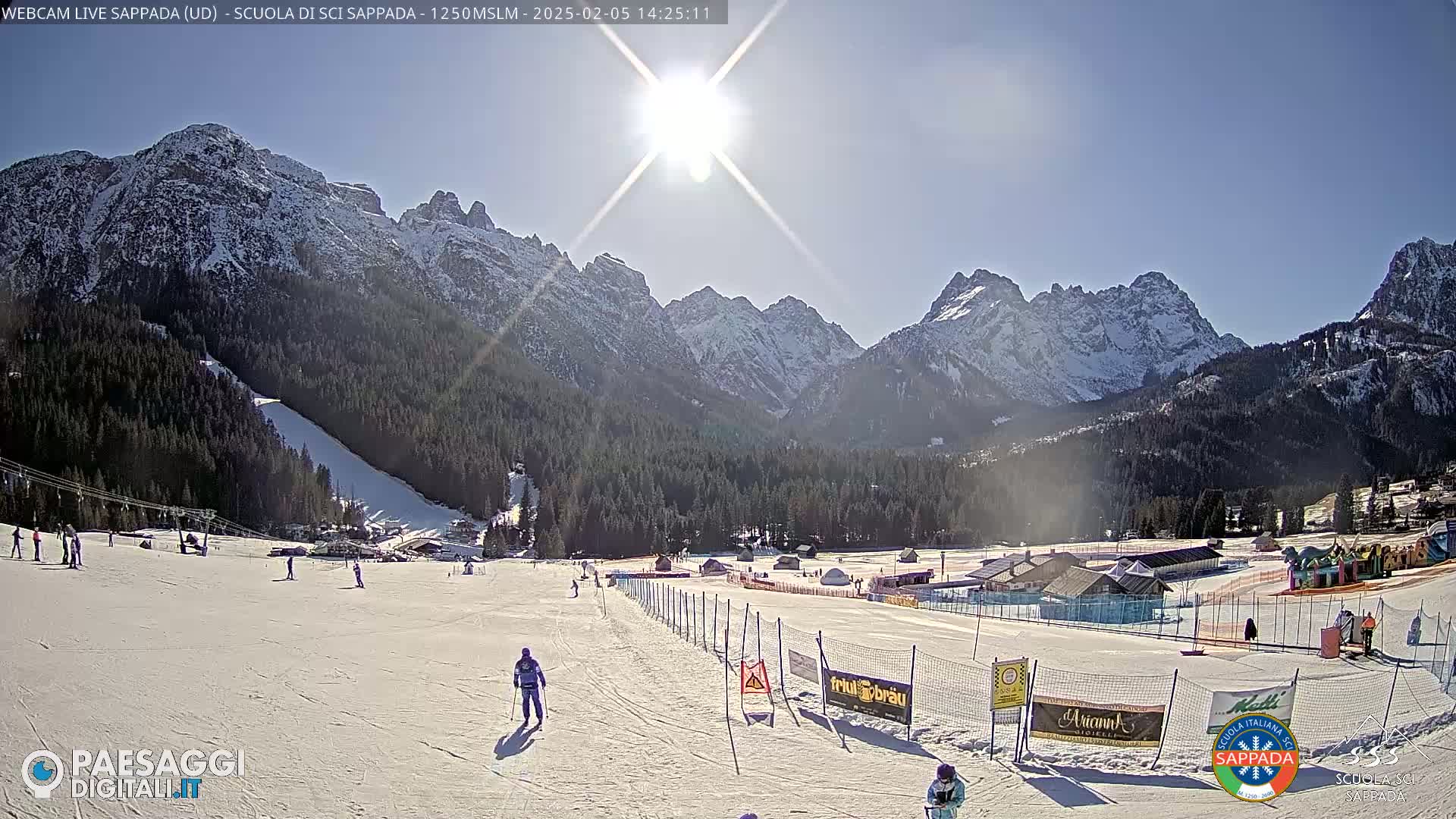 A sunny day at a snow-covered ski resort in the mountains, with several skiers visible on the slopes.