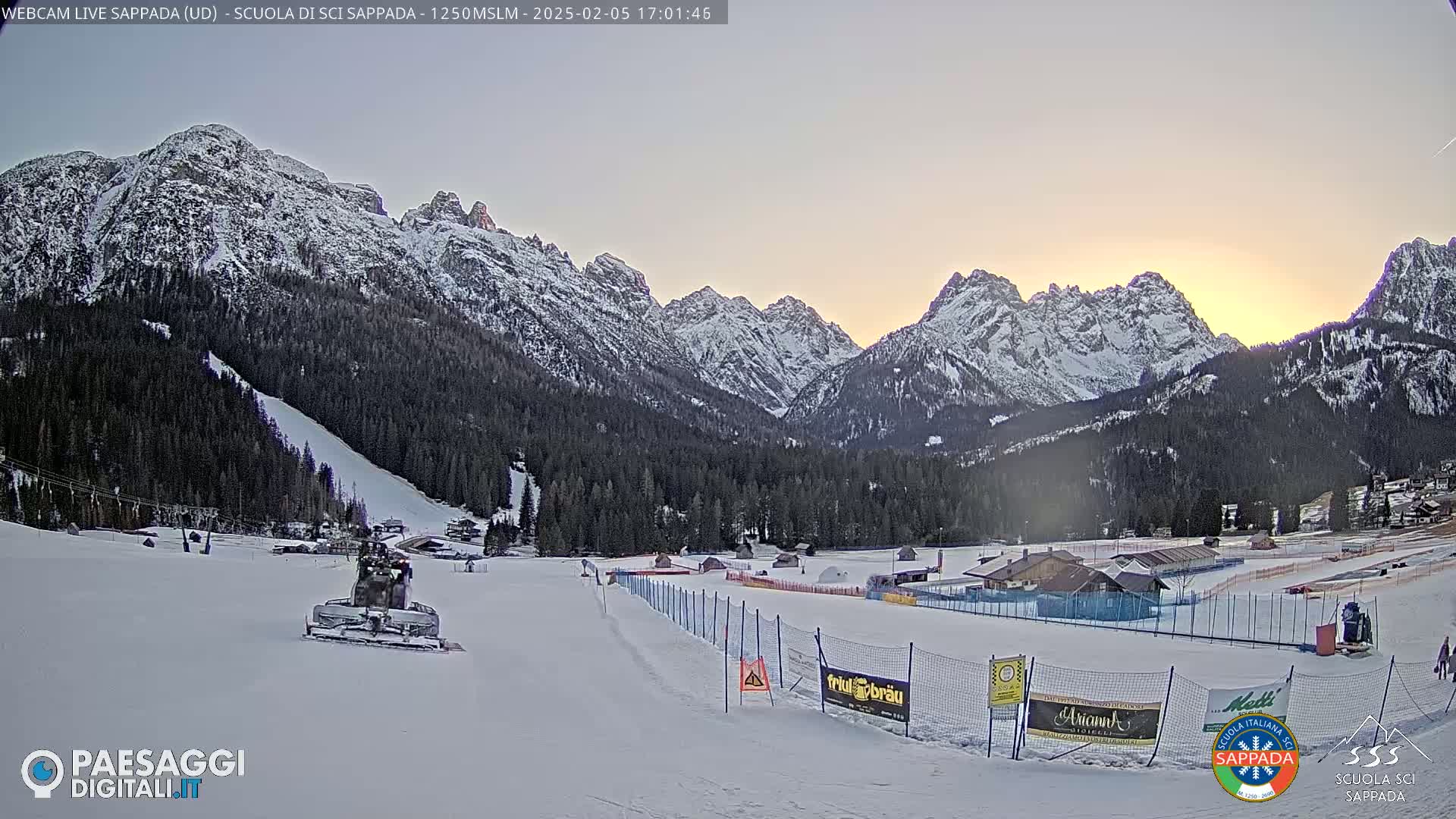 A snowcat grooms a snowy ski slope at sunset, with snow-covered mountains in the background.