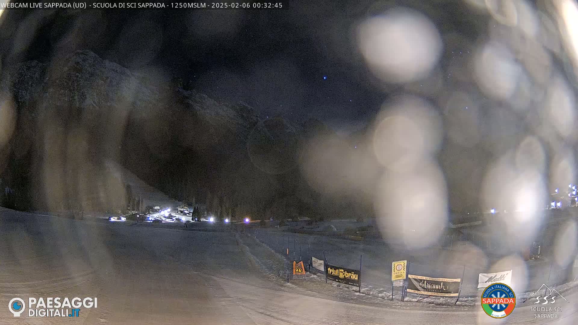 A nighttime, snowy scene shows a snow-covered slope with a few lights and blurry figures in the distance under a dark, starry sky.