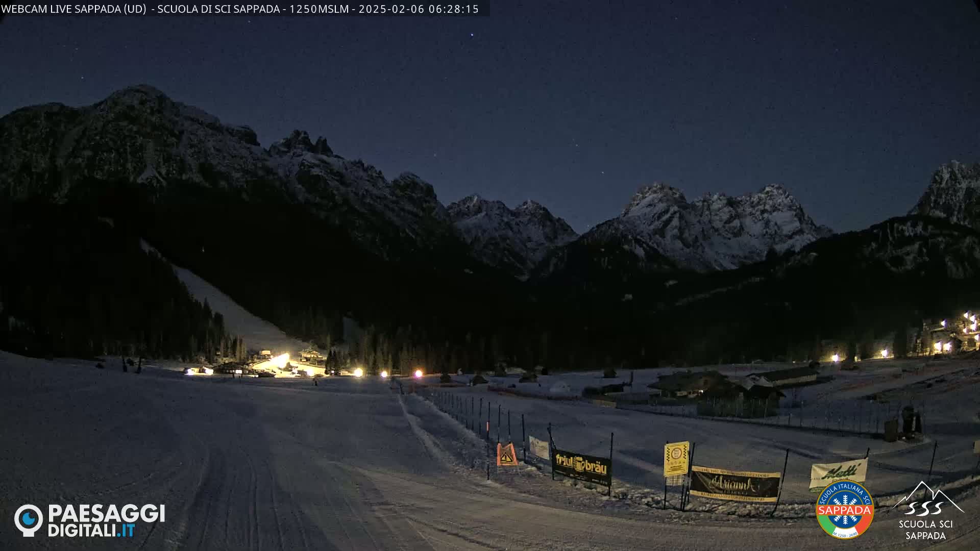 A nighttime view of a snow-covered ski slope at the base of dark, snow-capped mountains under a twilight sky.