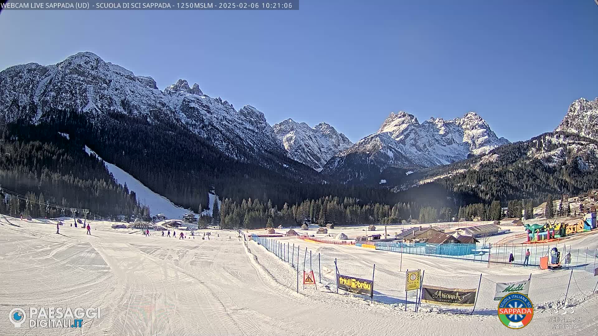 A snow-covered ski slope with several skiers, bordered by a fence, sits in front of a valley of snow-capped mountains under a clear blue sky.