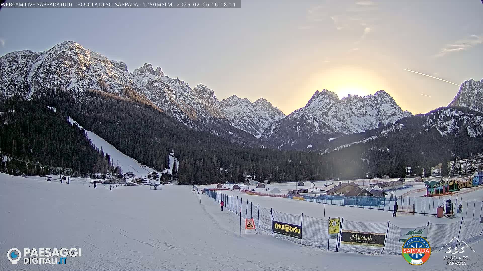 A snow-covered ski slope at sunset, with snow-capped mountains in the background.