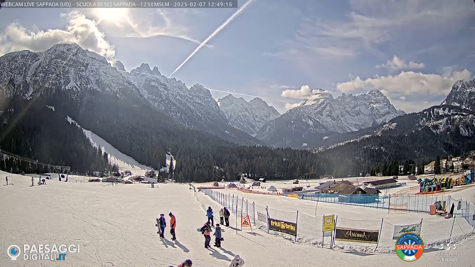 A snow-covered ski slope at the base of snow-capped mountains under a partly cloudy sky.