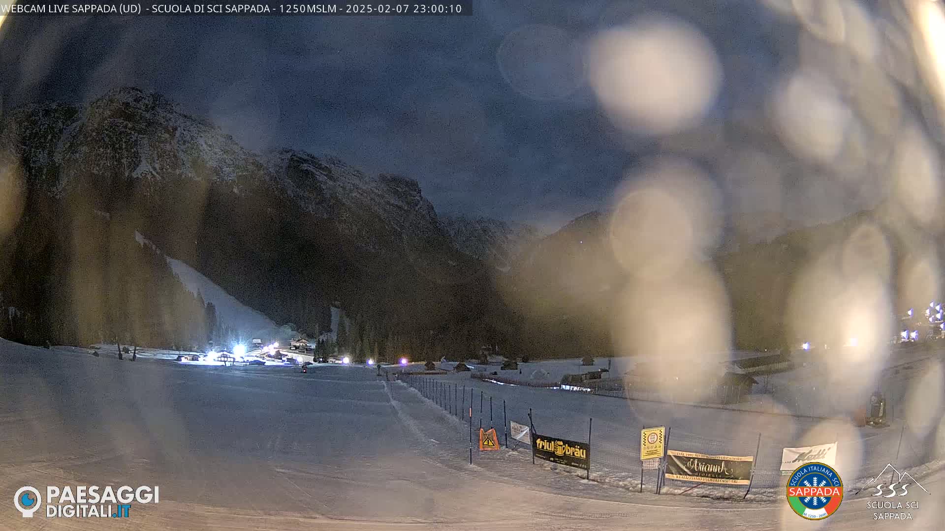 A nighttime view of a snow-covered ski slope at the base of a mountain under a dark, possibly cloudy sky.
