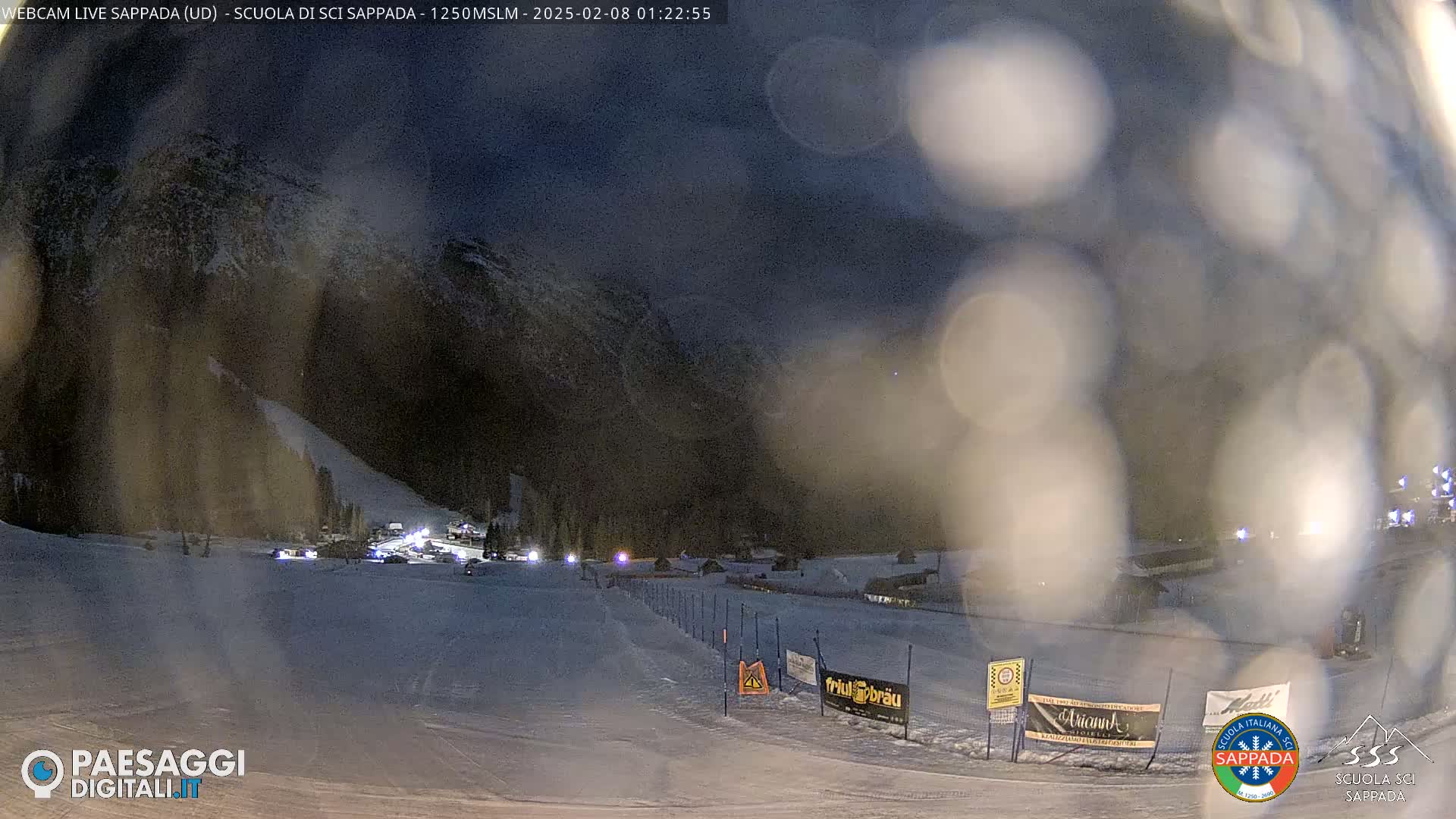 A nighttime view of a snow-covered ski slope with several lights visible in the distance, under a dark, possibly cloudy sky.