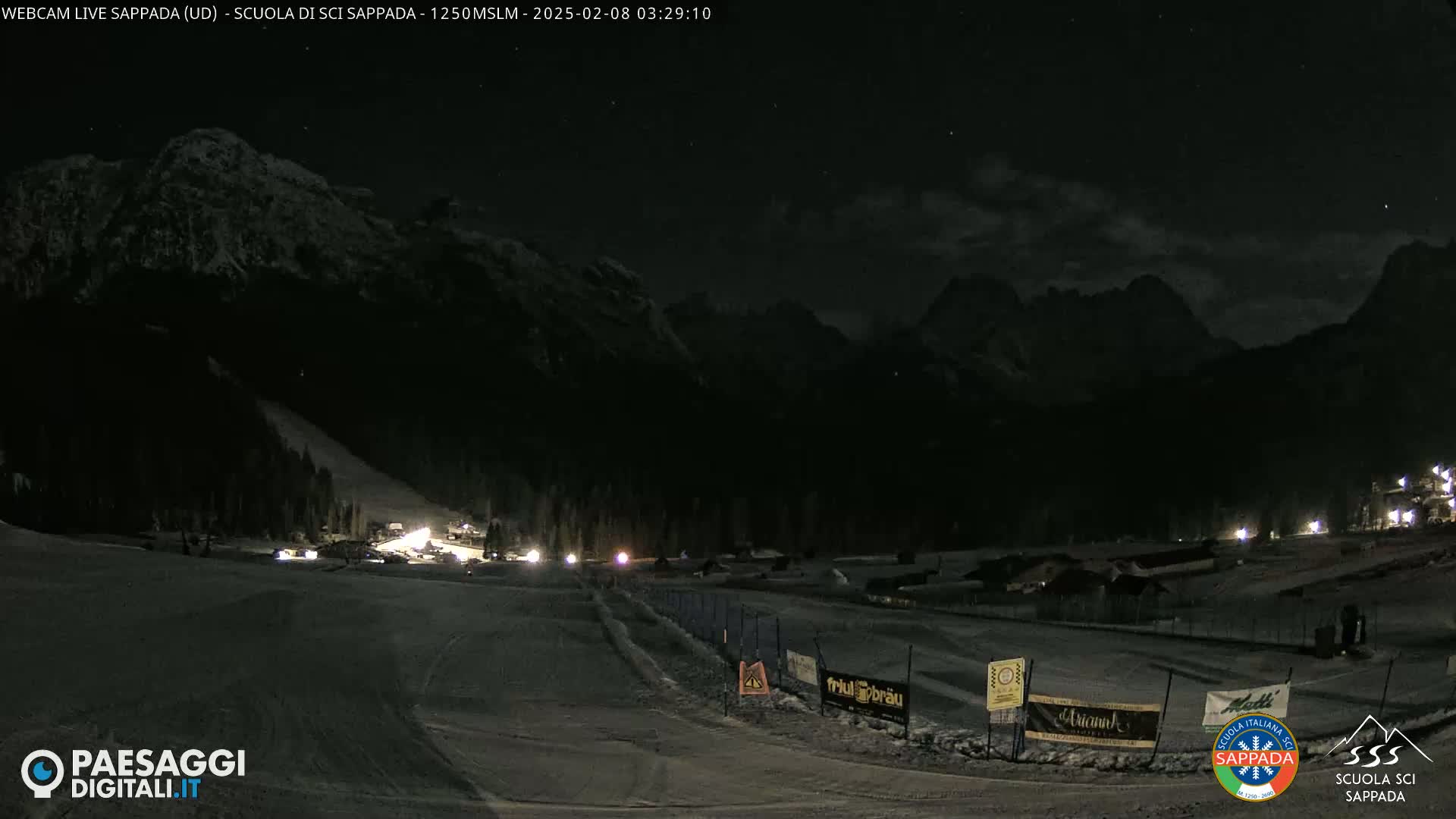 A nighttime view of a snow-covered ski slope and surrounding mountains under a partly cloudy sky.