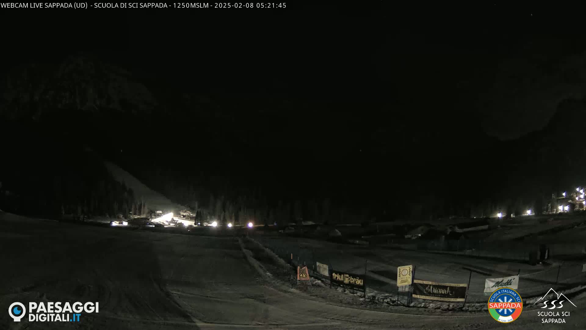 A dark, nighttime view of a snow-covered ski slope with sparsely lit buildings and vehicles in the distance.