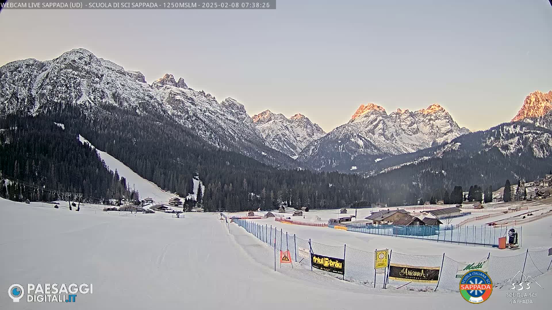 A snow-covered ski slope is situated in a valley below snow-capped mountains, bathed in the soft light of sunrise.